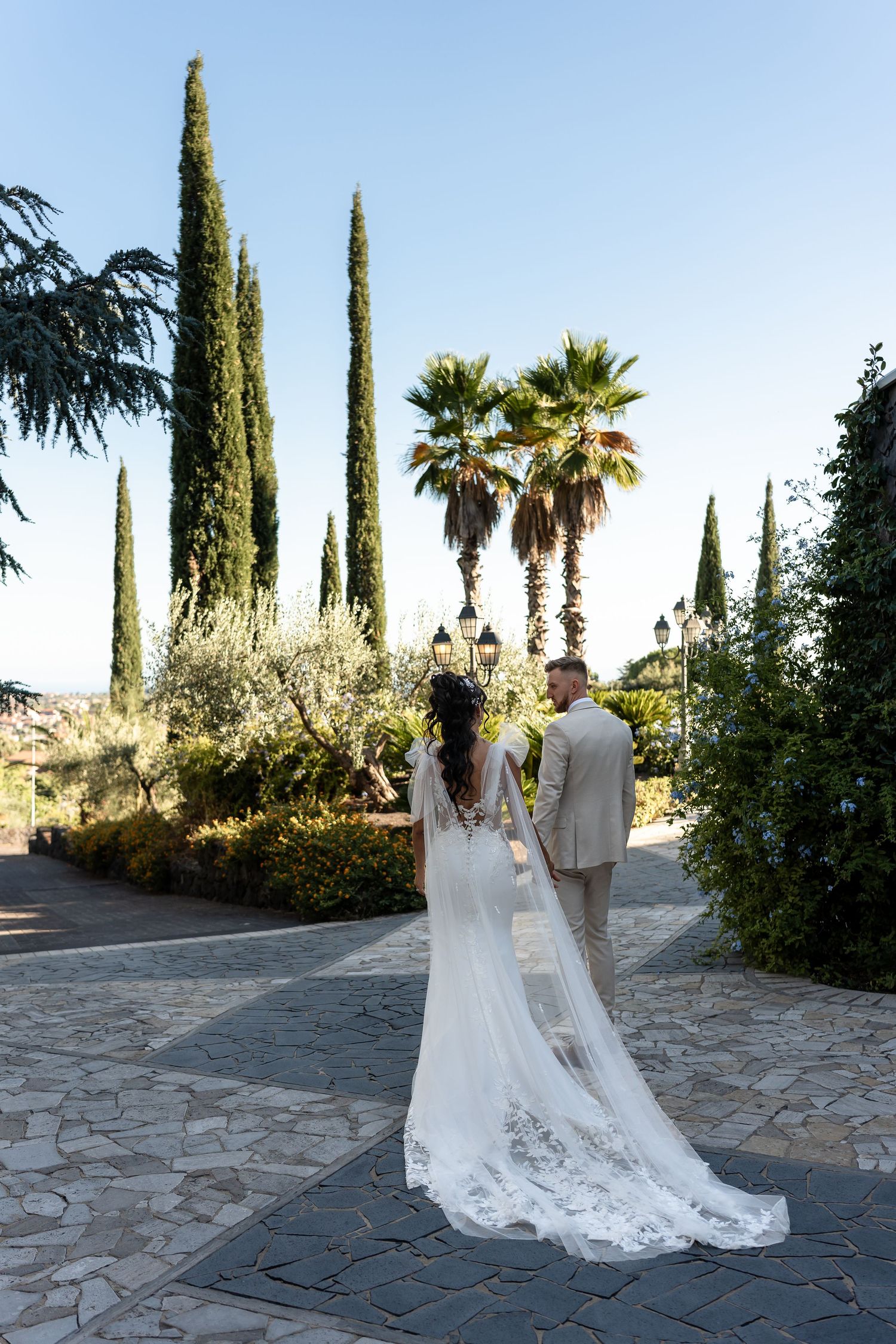 Bride in lace cathedral veil gown and groom walking among cypress and palm trees in Mediterranean garden.