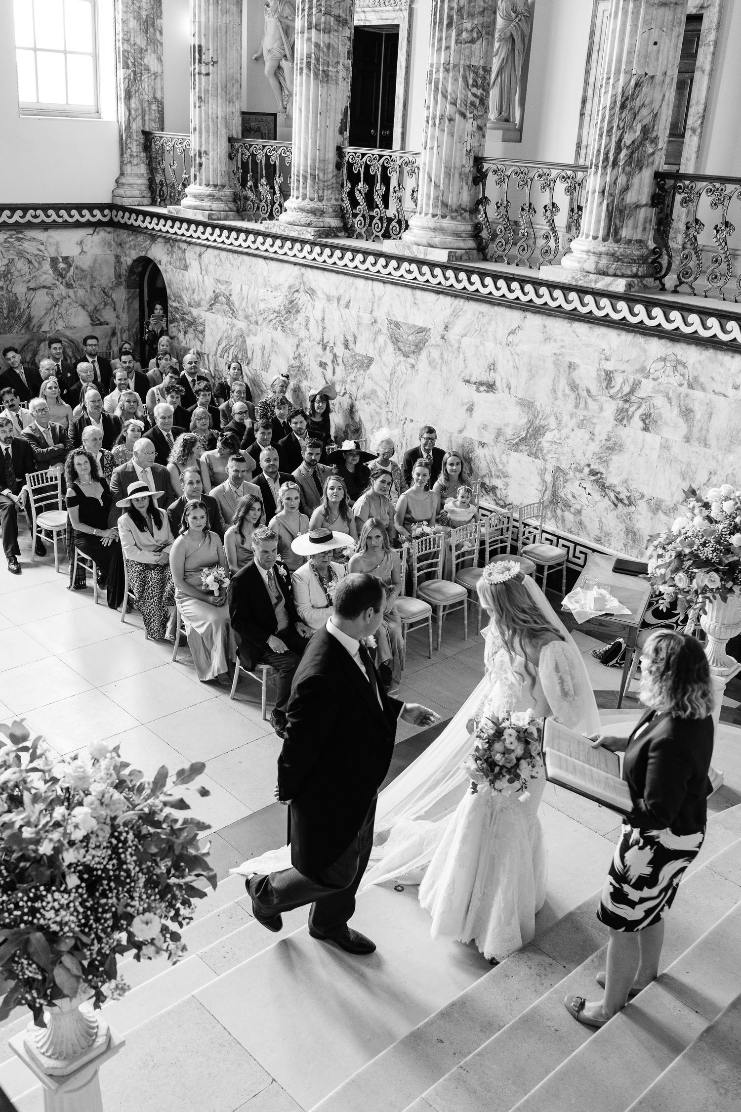 Black and white aerial view of a wedding ceremony in a grand marble hall with seated guests.