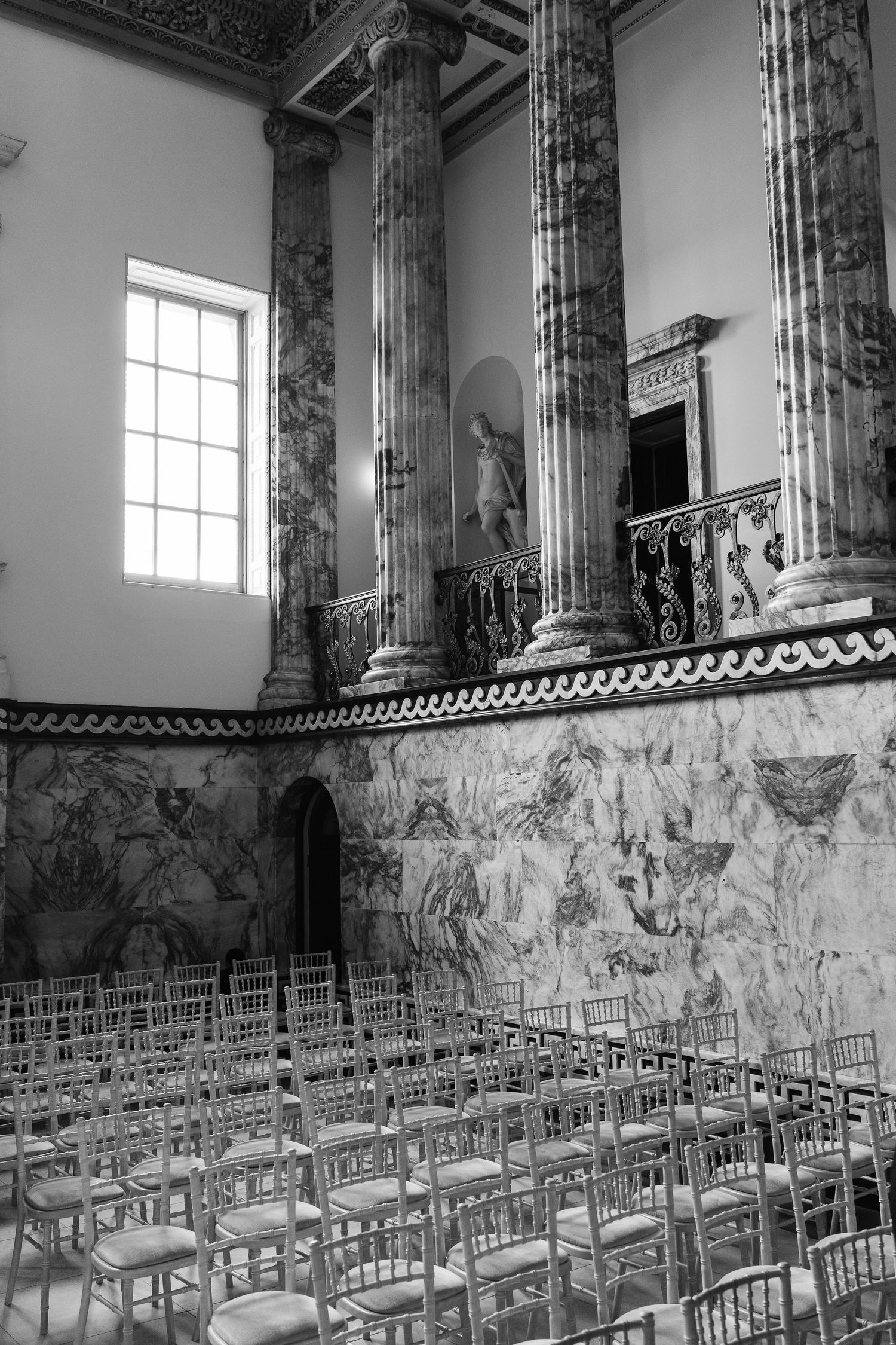 Black and white photo of ornate marble columns and painted walls in a grand hall with rows of elegant chairs.