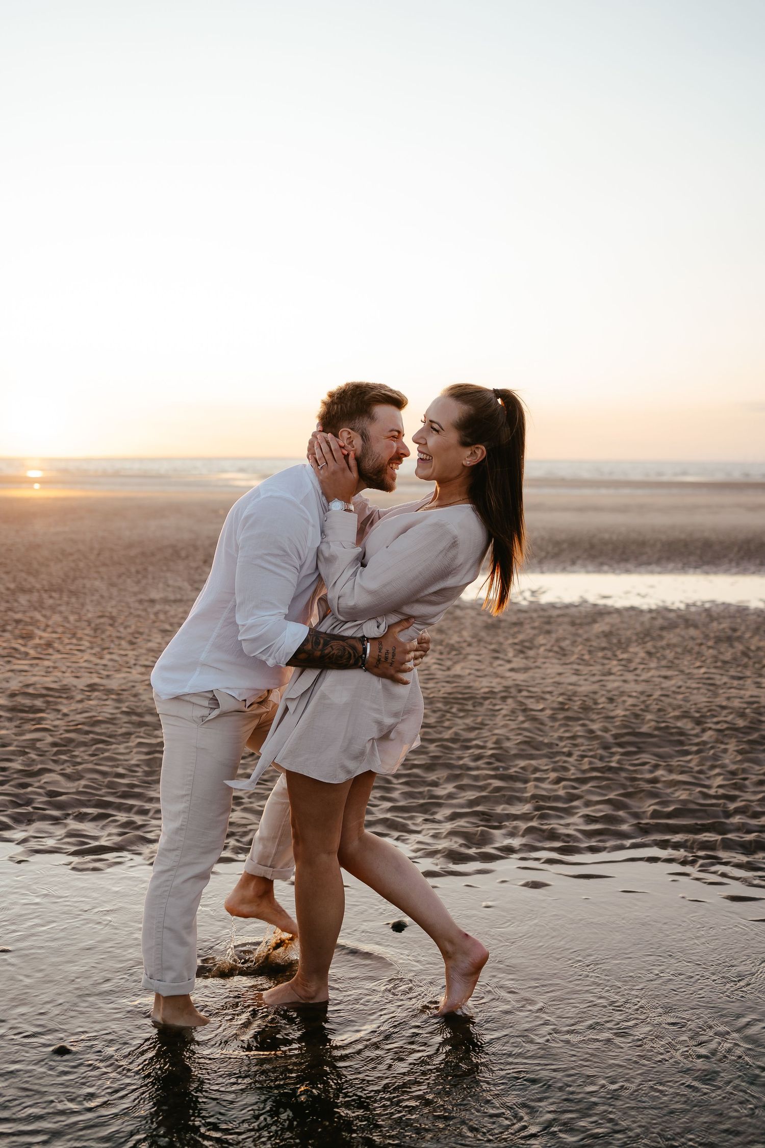 Laughing couple embracing on a reflective wet beach at sunset, dressed in casual white and beige outfits.