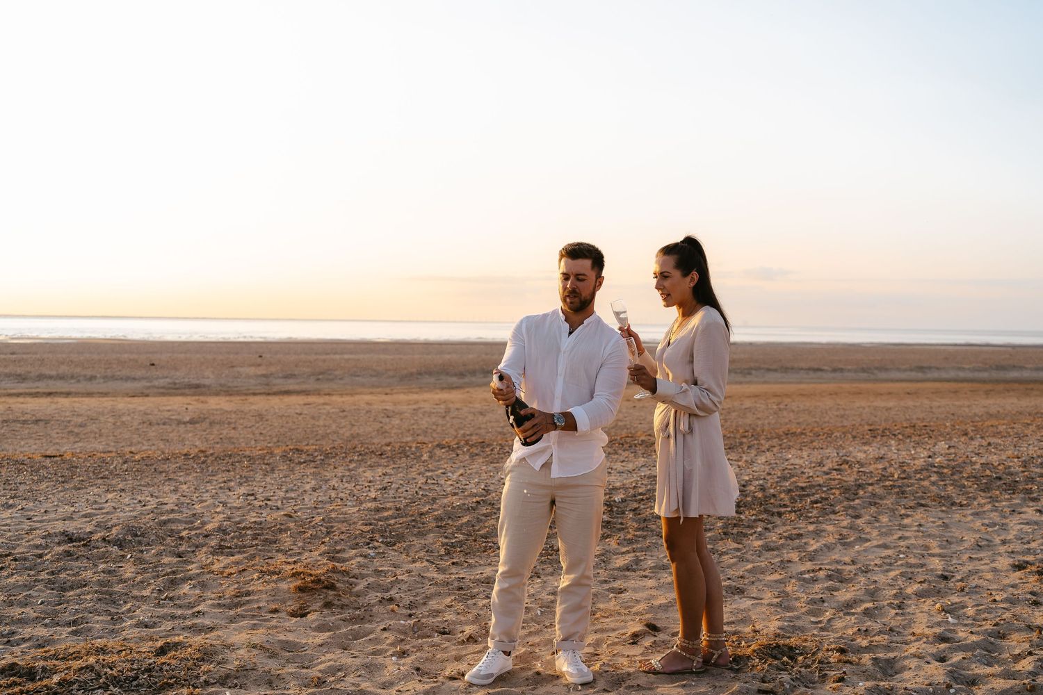 Couple standing on a sandy beach at sunset, sharing champagne glasses in matching white outfits.