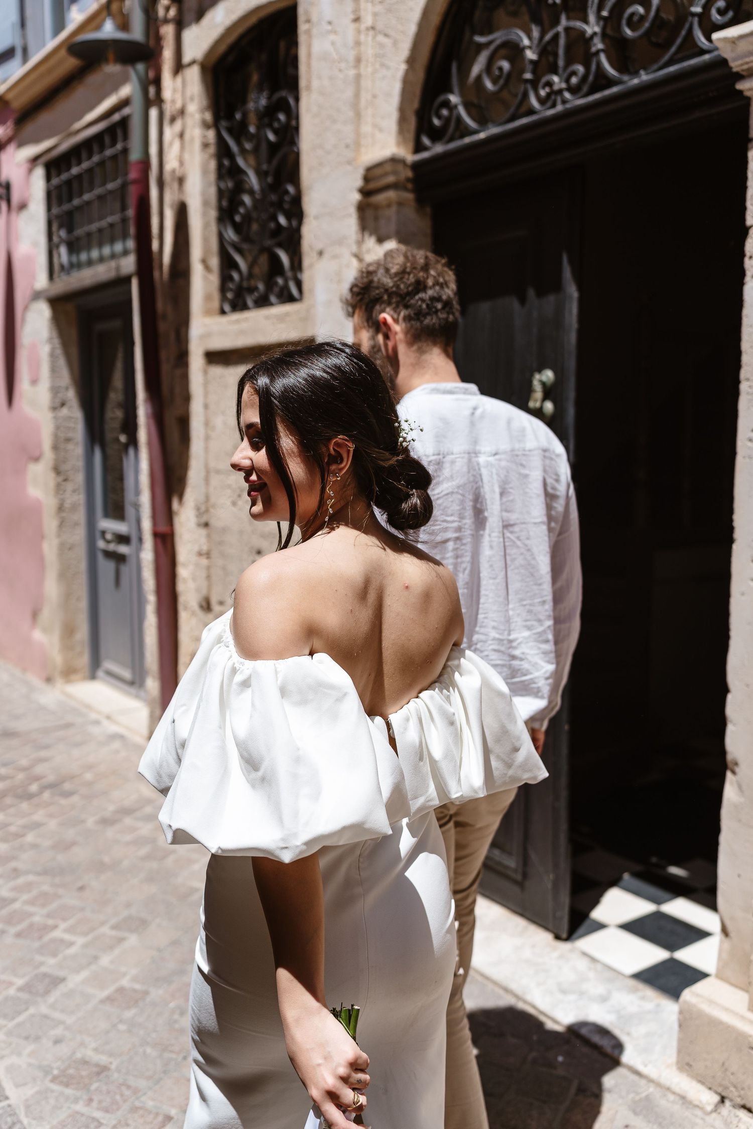 Smiling bride in off-shoulder white dress walking with groom through narrow European old town street.