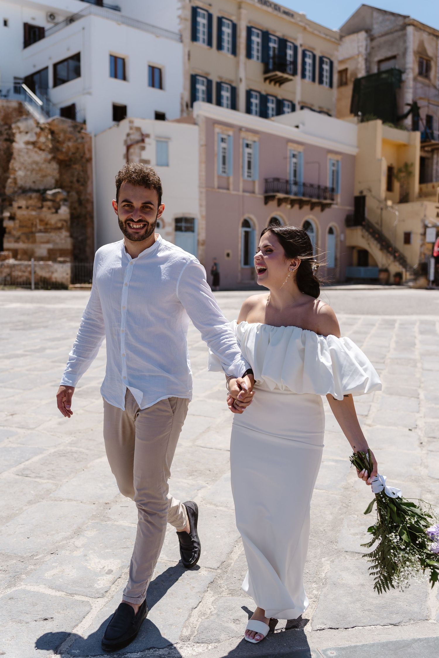 Happy bride and groom laughing while strolling hand-in-hand through a sunlit Mediterranean town square.