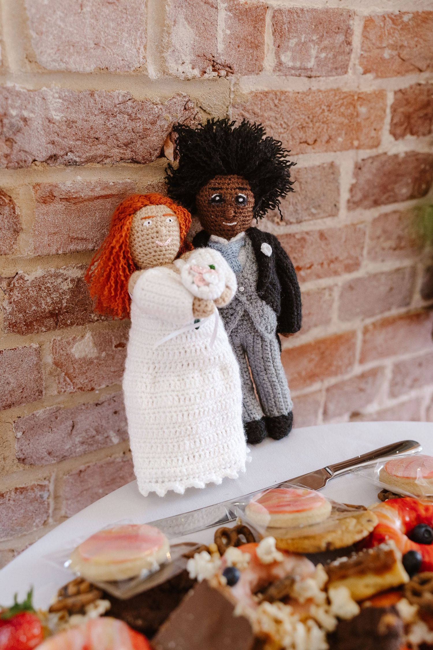 Handmade crochet bride and groom wedding cake toppers leaning against a rustic brick wall.