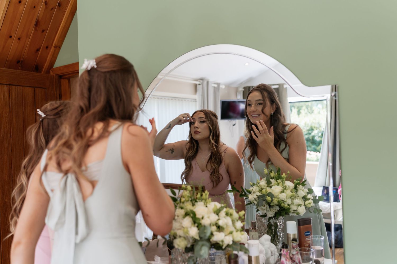 Two bridesmaids admiring their wedding morning makeup looks together in a large ornate mirror.