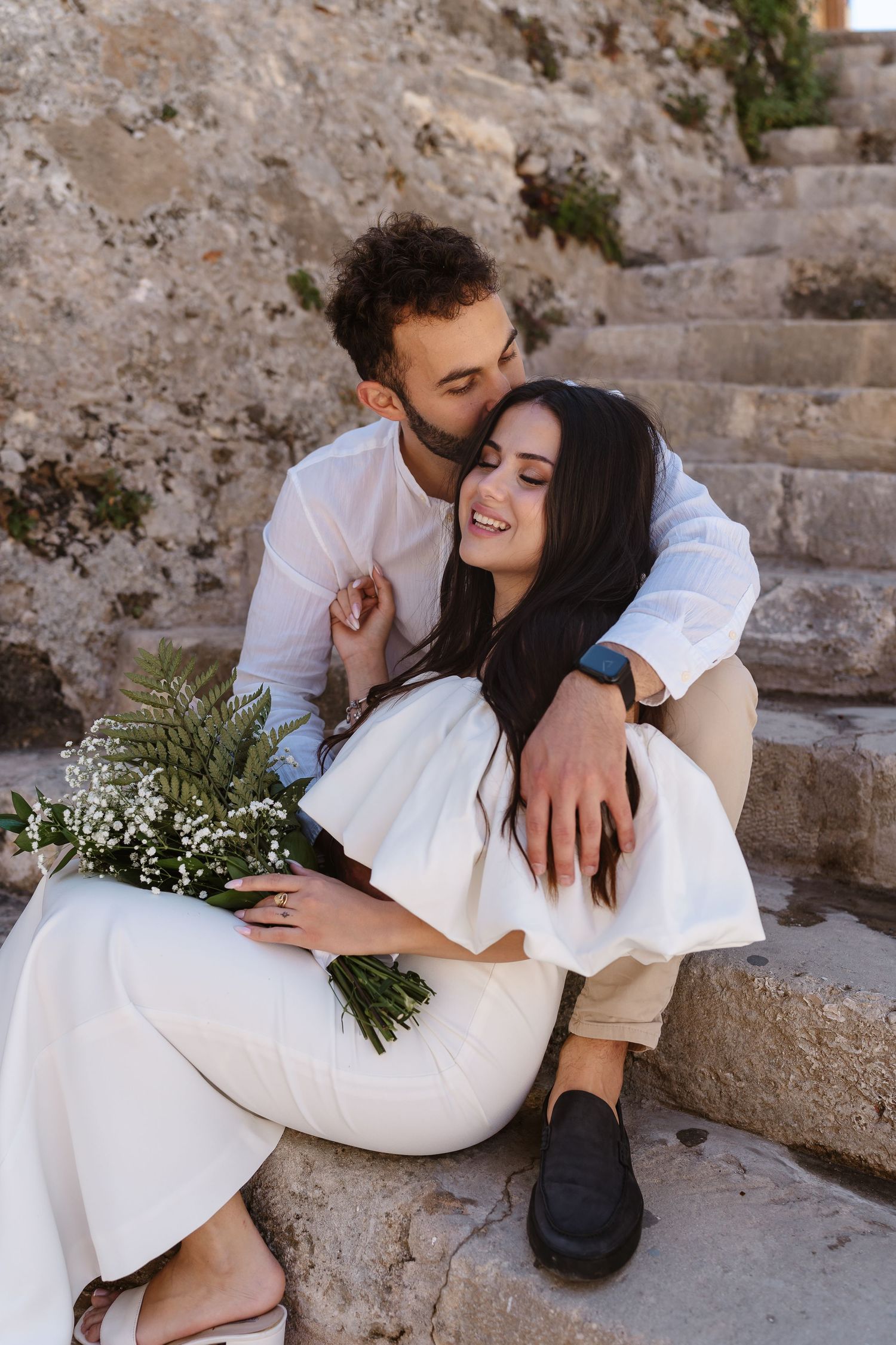 Groom kissing smiling bride on ancient stone steps while she holds white wildflower wedding bouquet.