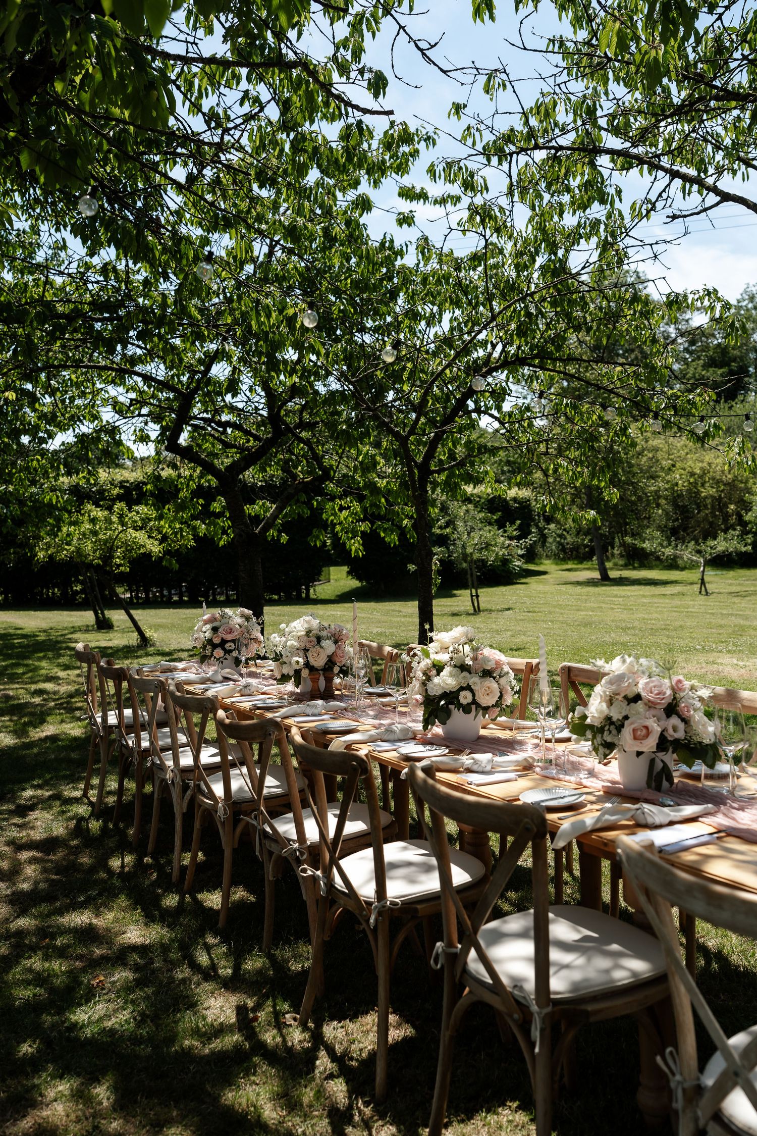 Long outdoor wedding reception table set with floral centerpieces under leafy green trees in a garden.
