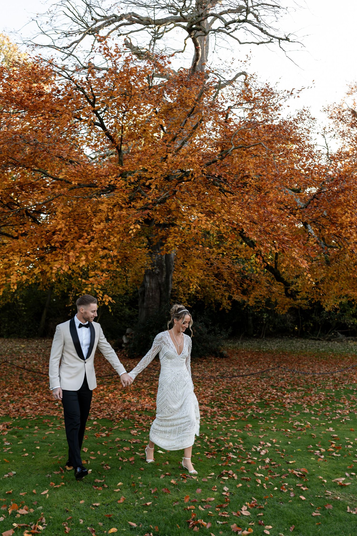 A bride and groom walk hand-in-hand beneath a stunning autumn tree with orange foliage.