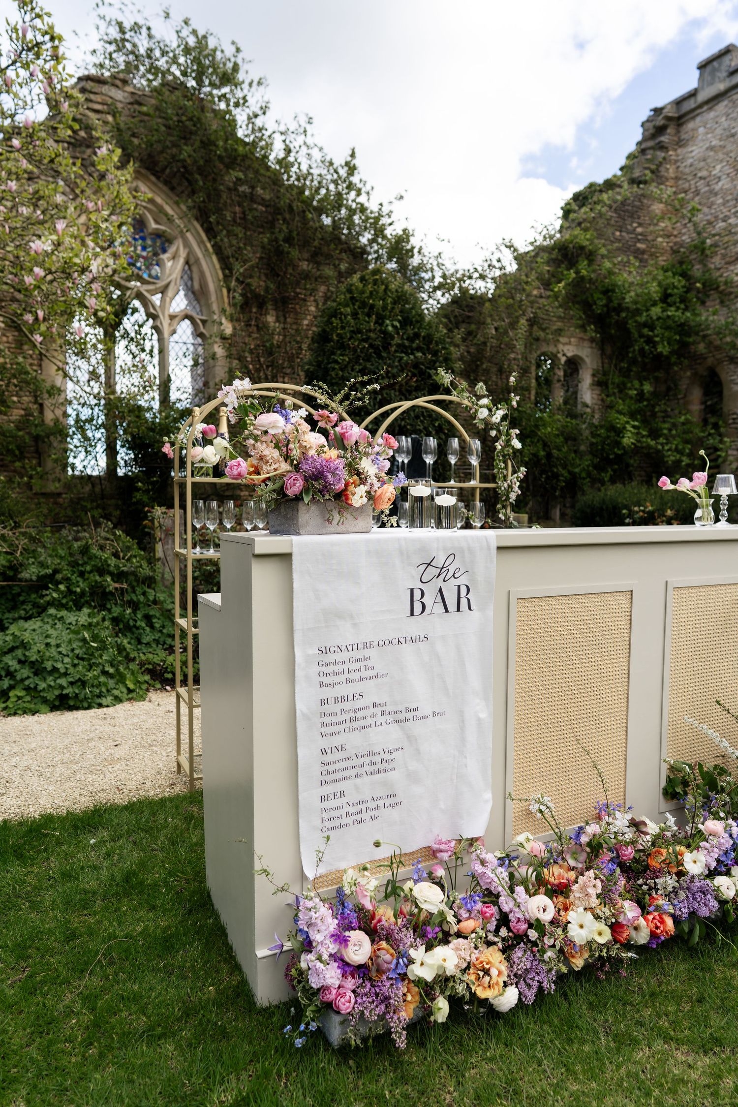 Outdoor wedding bar decorated with colorful floral arrangements set against a rustic stone ruin backdrop.