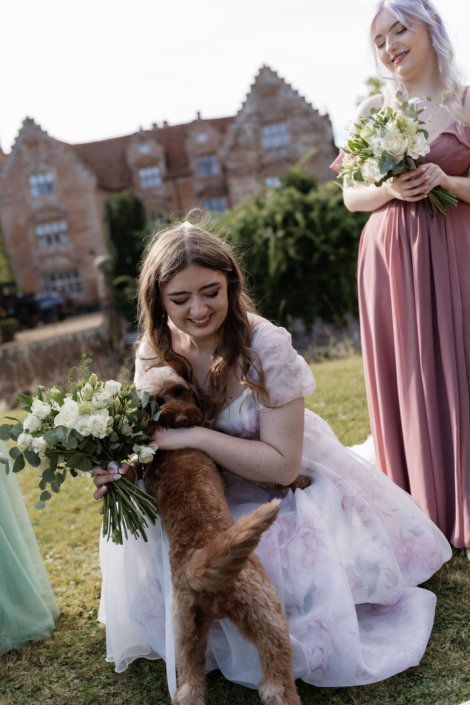Smiling bride in floral gown crouches to pet a golden dog at her wedding.