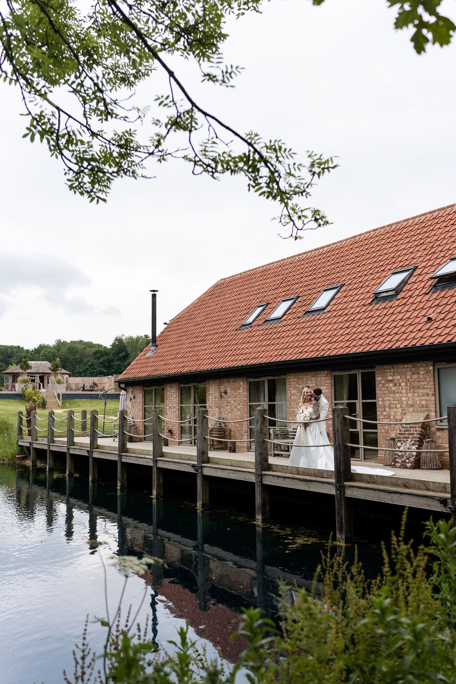 Bride and groom embrace on waterside deck of rustic brick wedding venue building.
