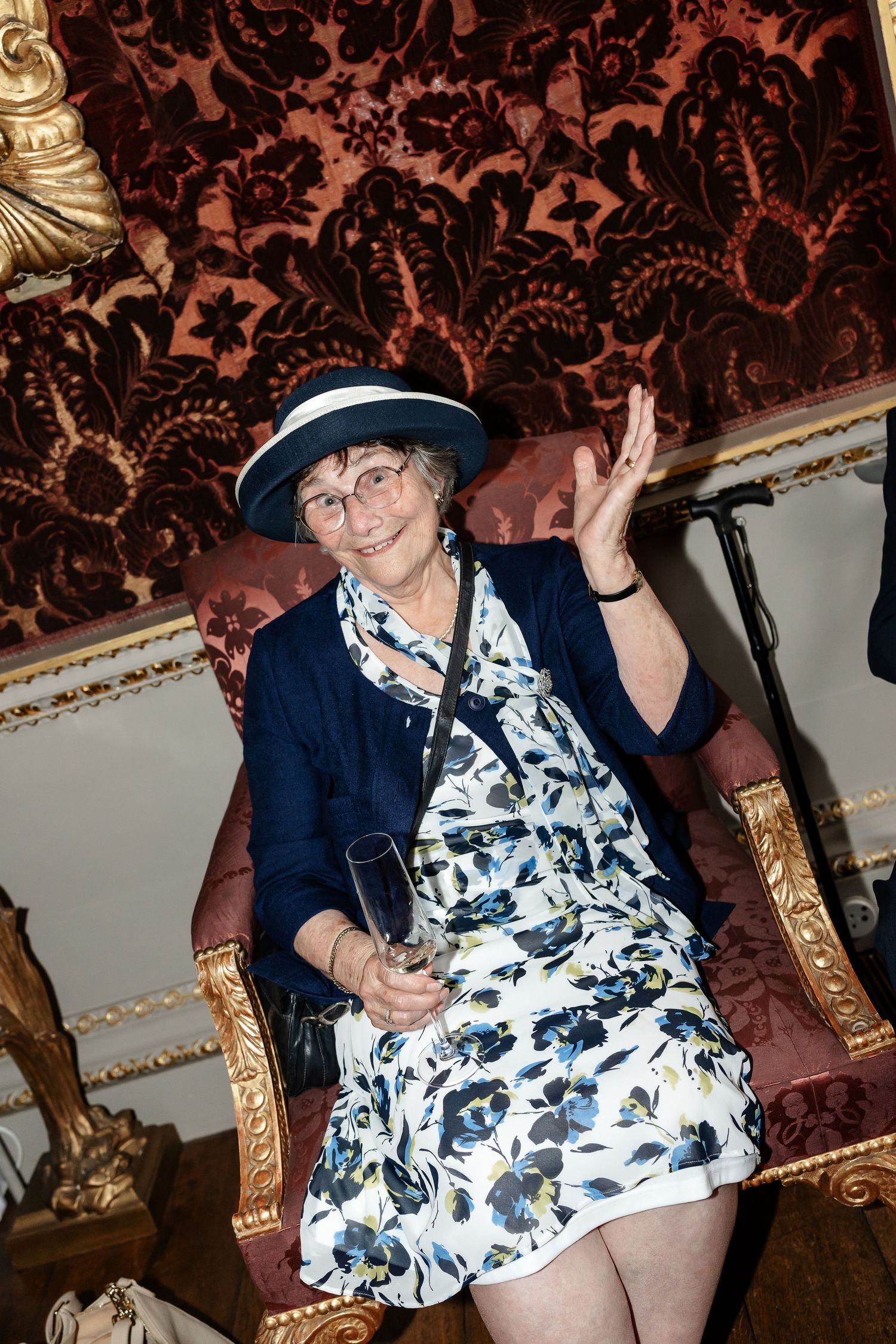 Elderly woman in floral dress and white hat waves while seated on ornate gold chair at wedding.