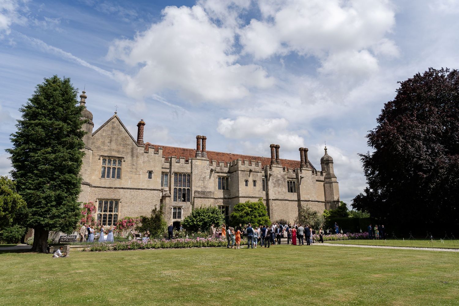 Wedding guests gather on the lawn of a grand historic English manor under a bright blue sky.