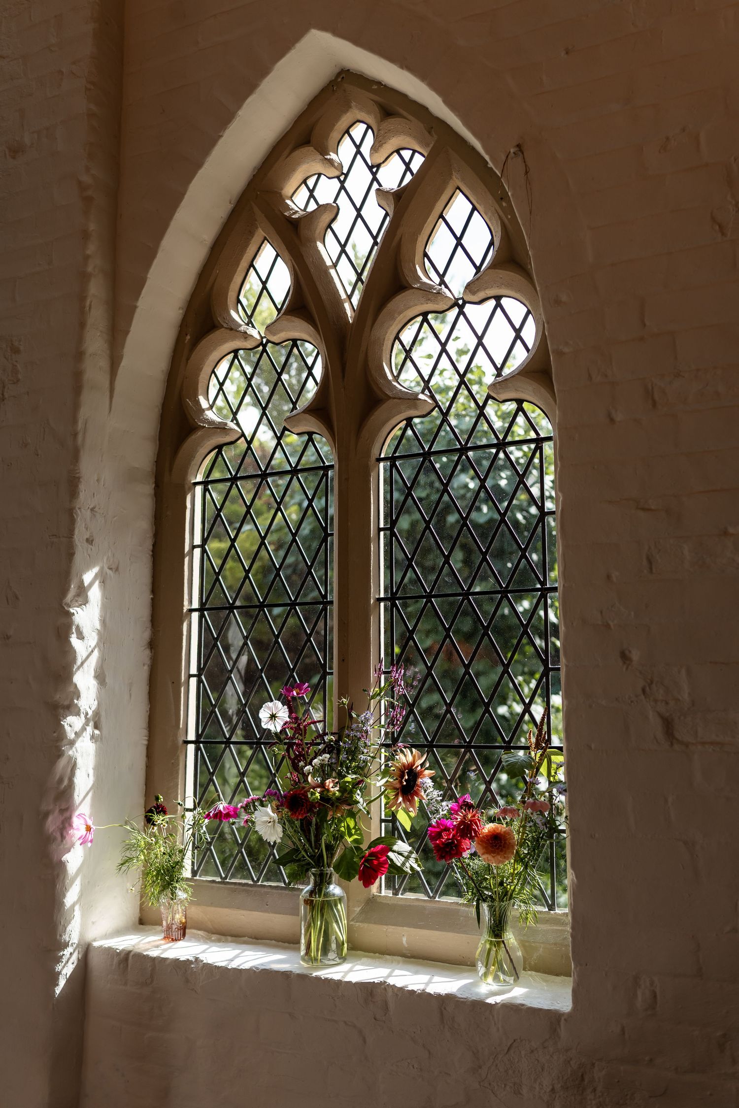 Colorful wildflower bouquets in glass vases on the sill of a Gothic arched leaded church window.