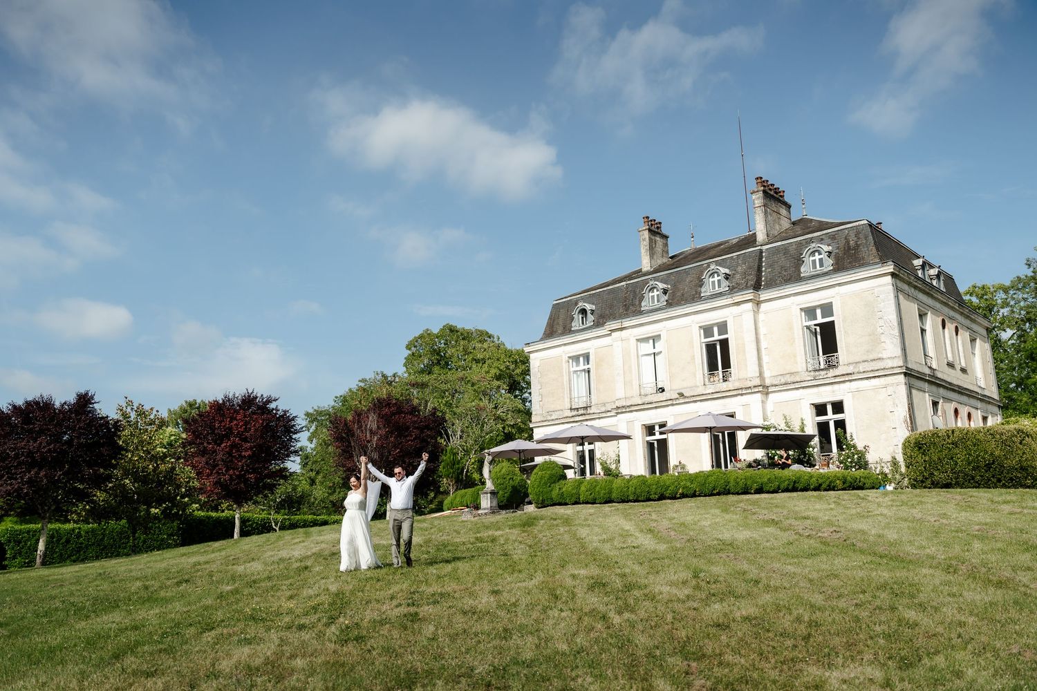 Bride and groom celebrating on vast green lawn before grand French chateau mansion under blue sky.