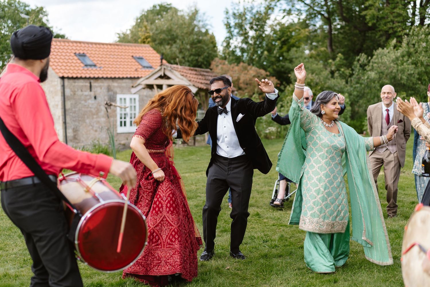Bride in red lehenga and guests dancing outdoors to live dhol drumming at Indian wedding.