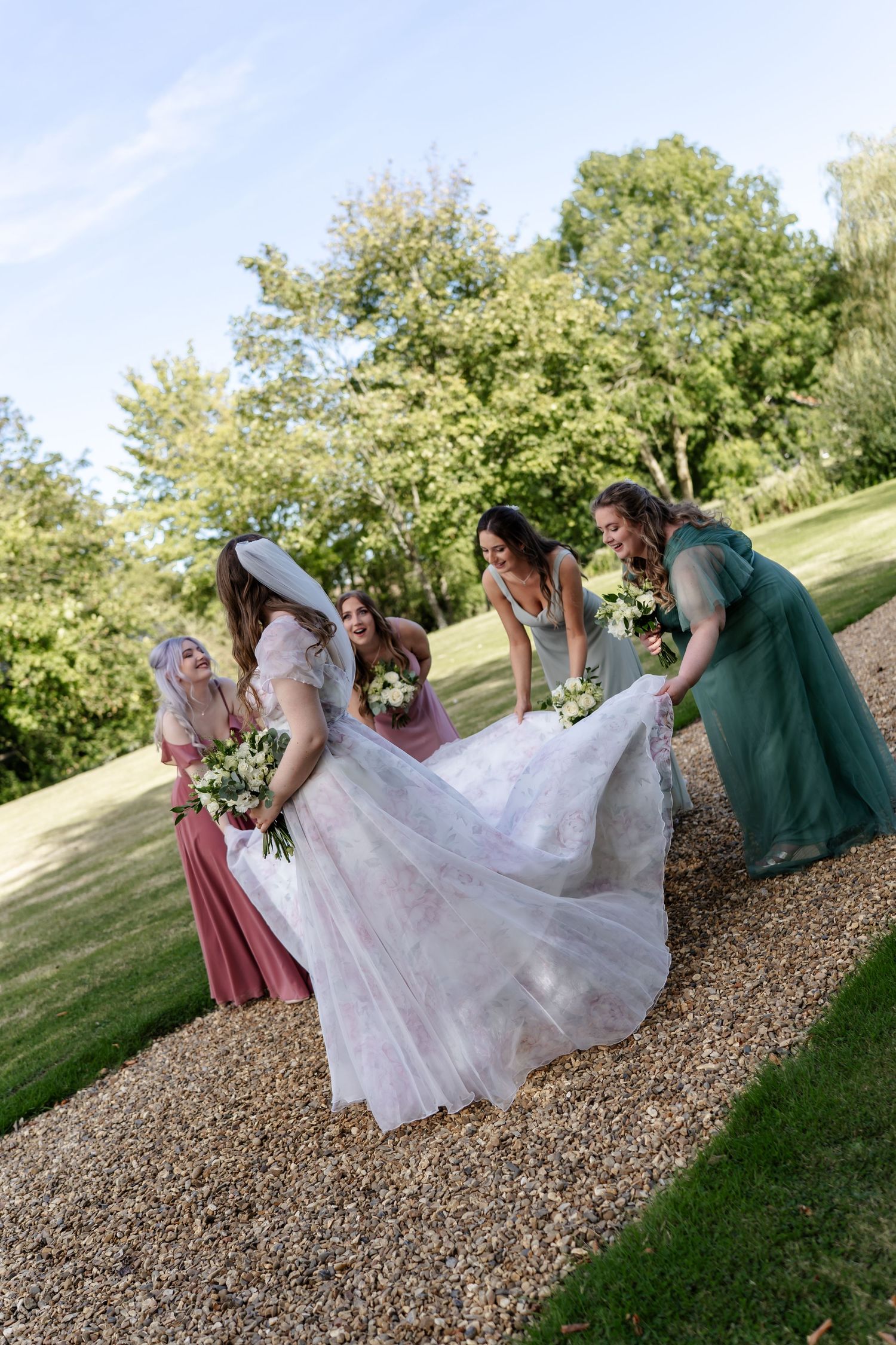 Bridesmaids in pink and green gowns lift bride's flowing tulle dress outdoors on gravel path.