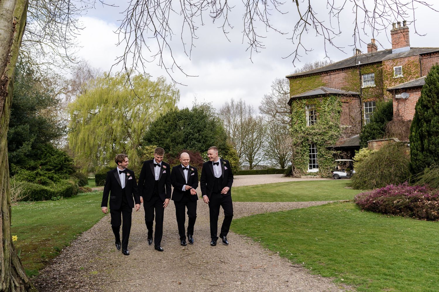 Four groomsmen in black tuxedos walking along a garden path beside an elegant English country manor.