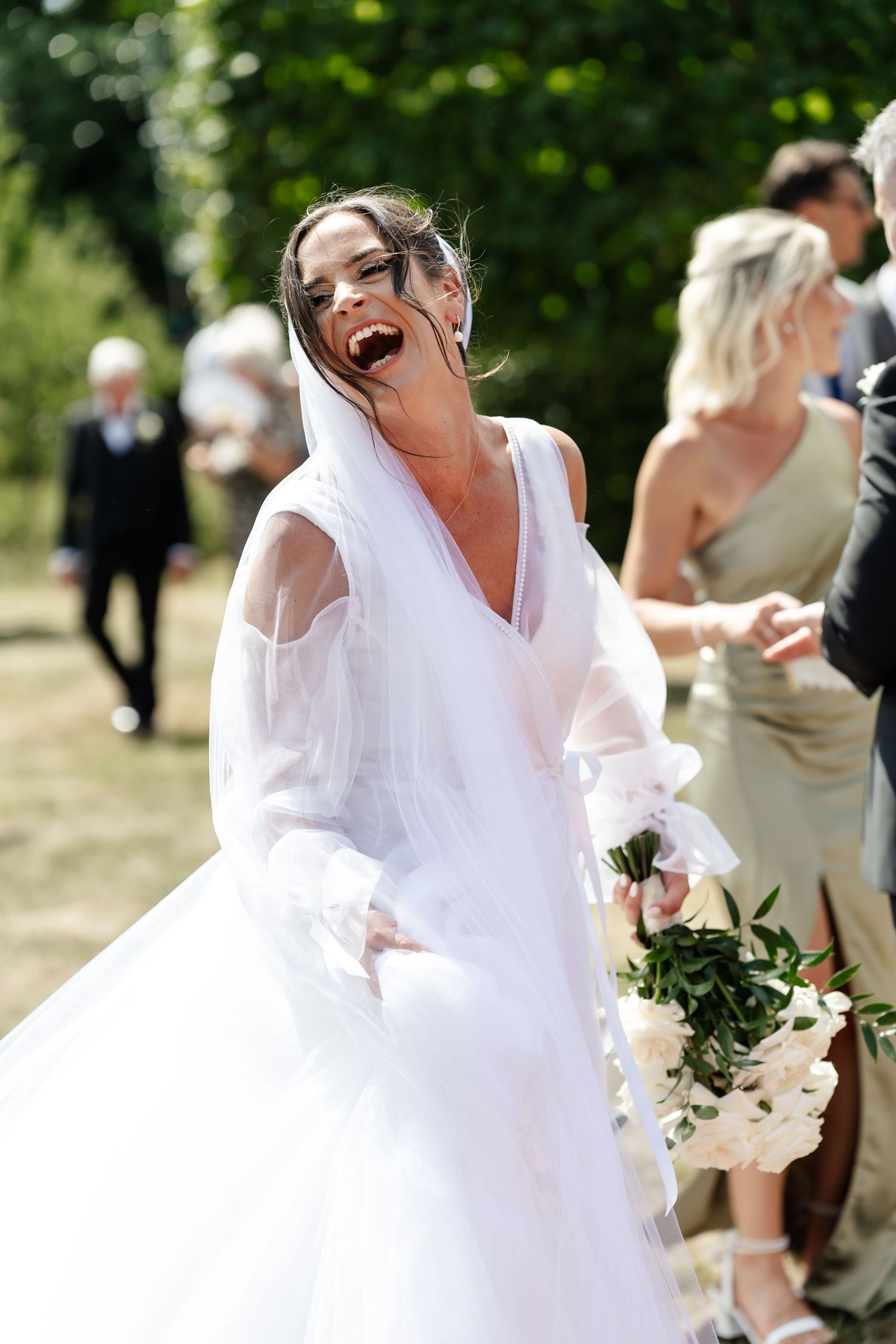 Joyful bride laughing in a white gown and veil while holding a bouquet outdoors.