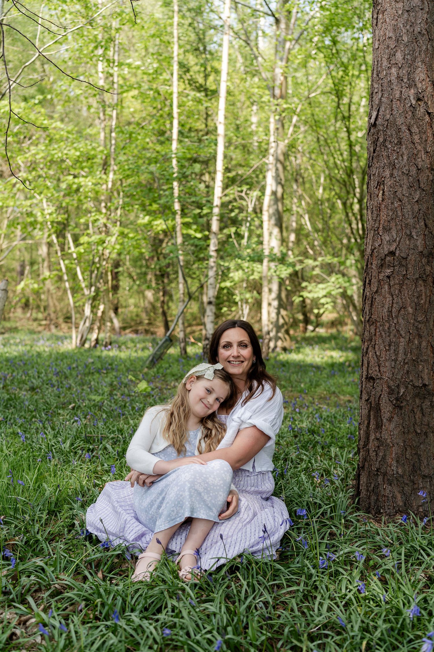 Mother and daughter sit together among bluebells in a sunlit woodland forest setting.