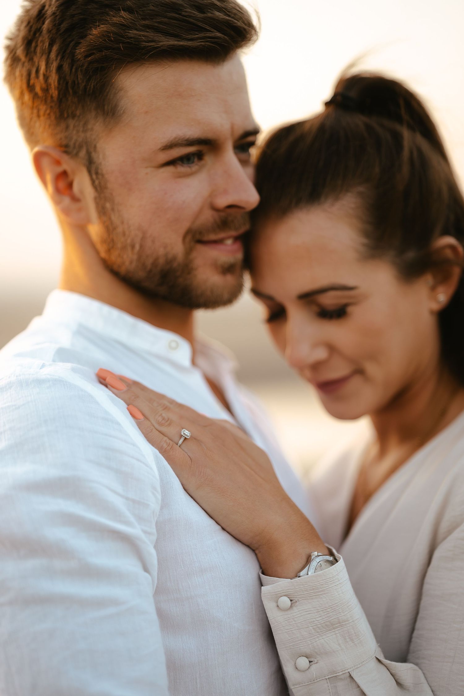 Close-up of couple embracing at golden hour, woman resting head on mans shoulder showing engagement ring.
