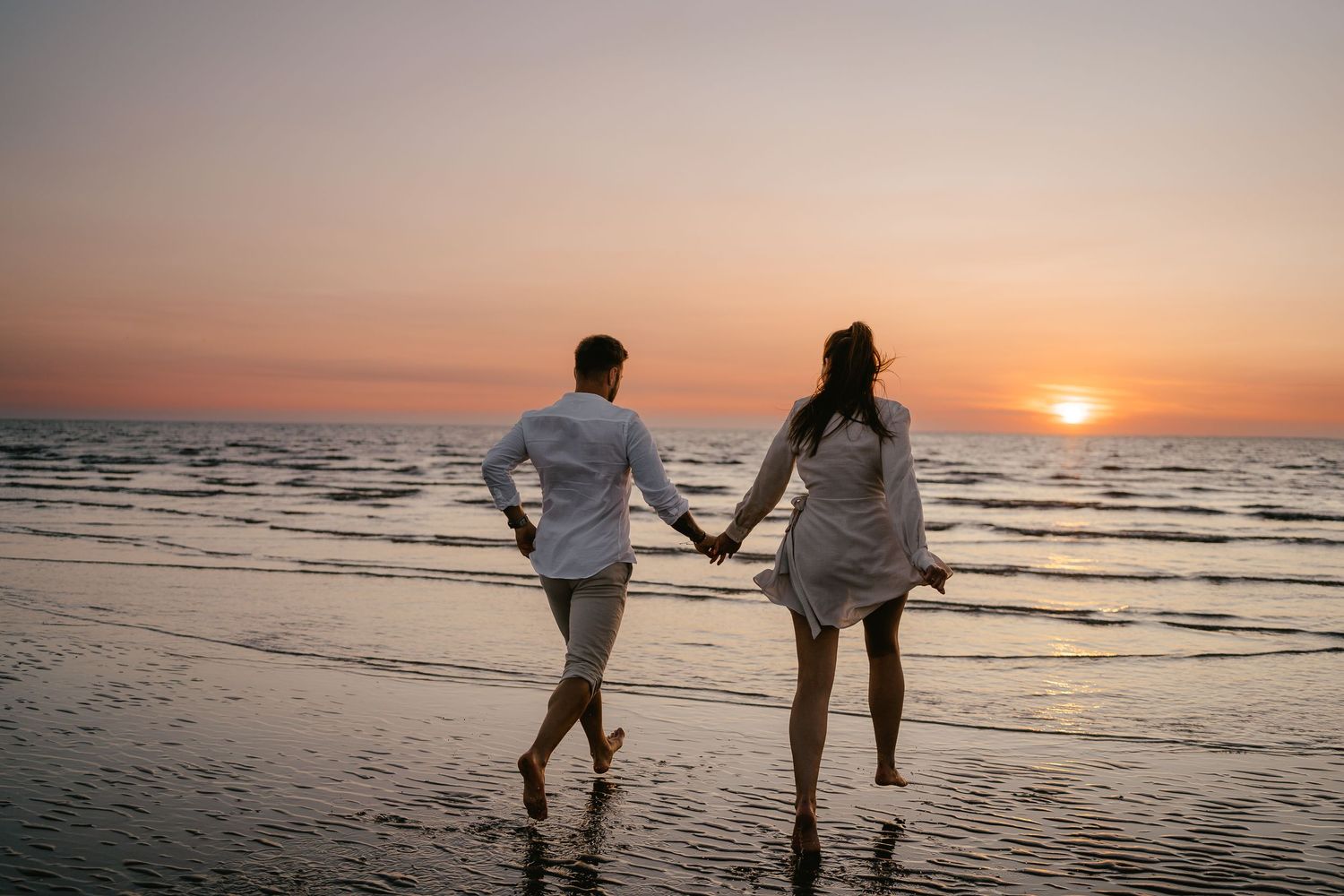 A couple holding hands while walking along a reflective shoreline, silhouetted against a colorful sunset.