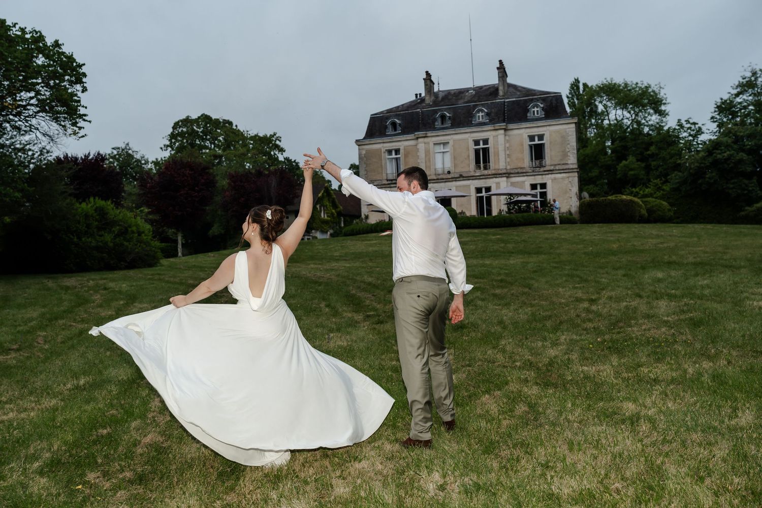 Bride and groom dancing joyfully on green lawn with elegant French chateau in background.