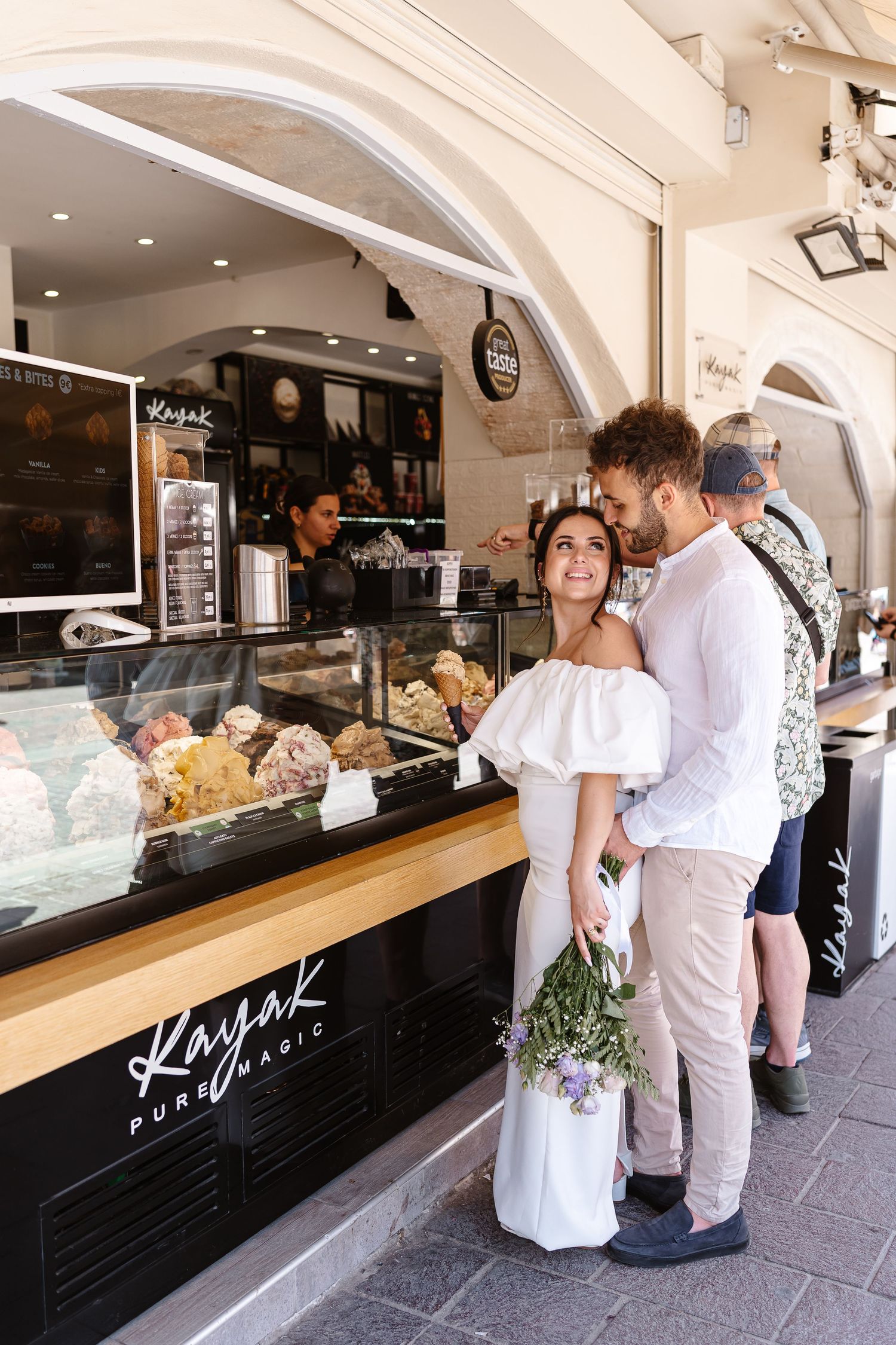 Couple in wedding attire ordering gelato at Kayak Pure Magic ice cream shop on a sunny day.