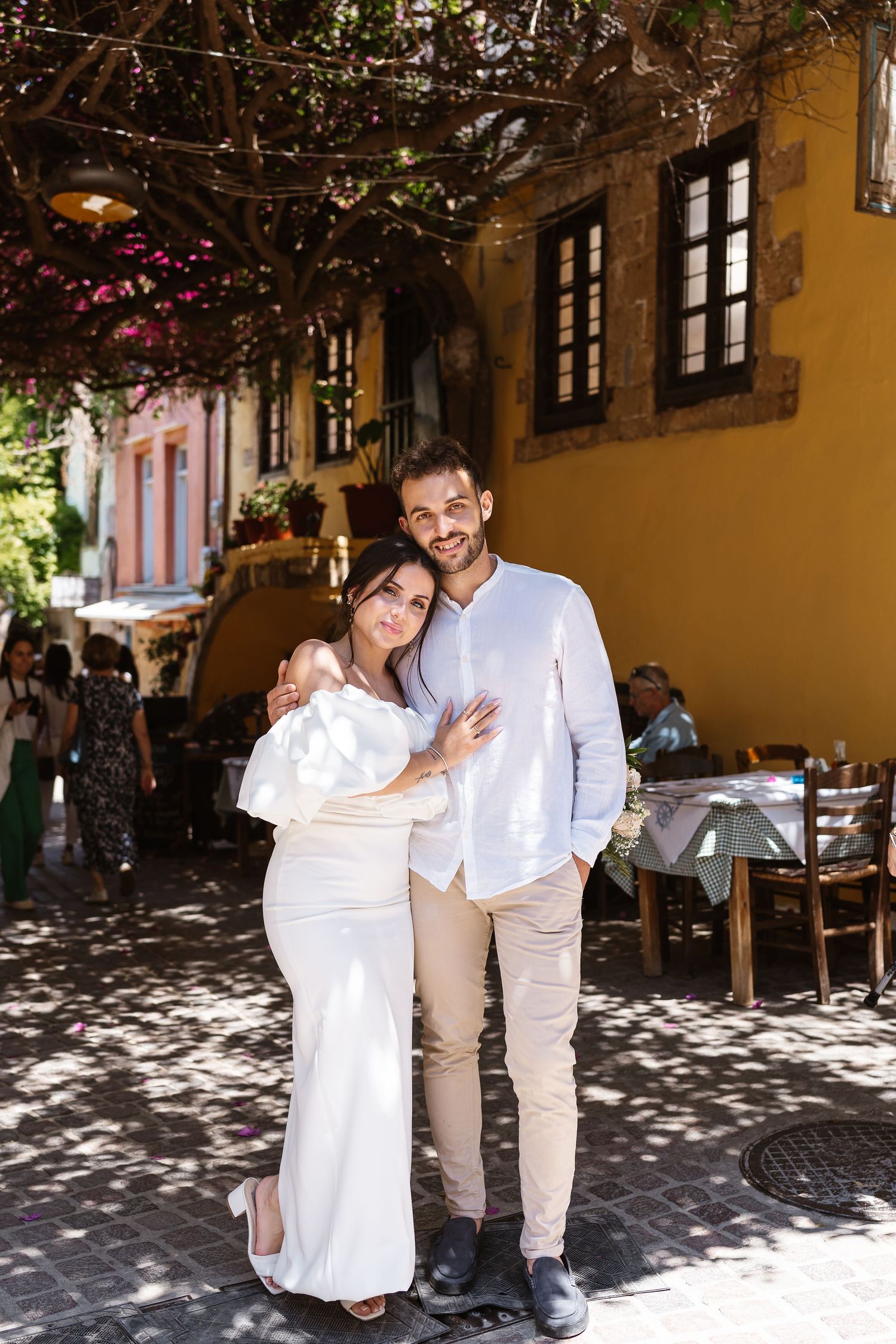 Elegant couple embracing under vine-covered pergola outside yellow Mediterranean building, groom smiling warmly.