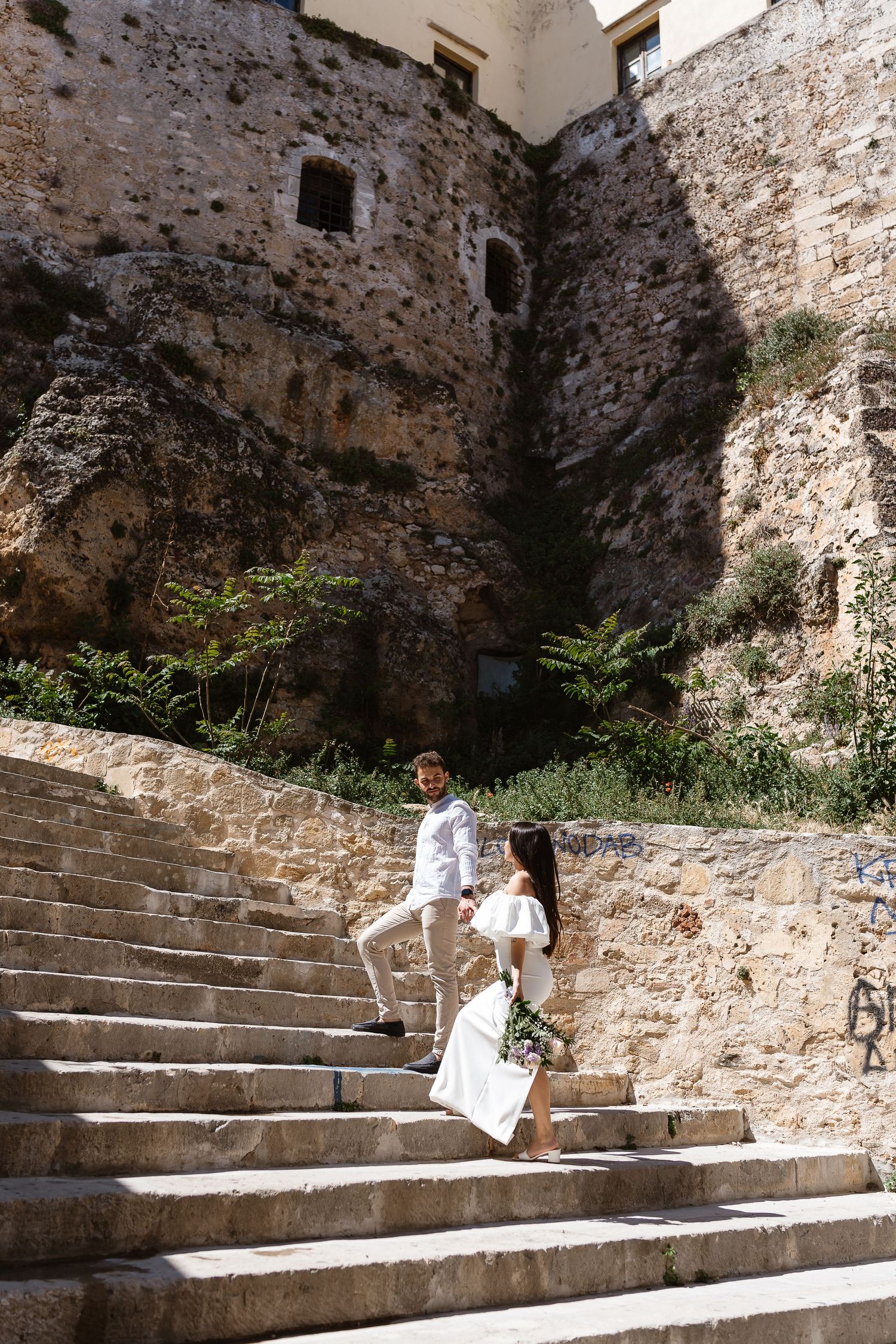 Couple climbing ancient stone staircase holding hands beside historic Mediterranean fortress wall.