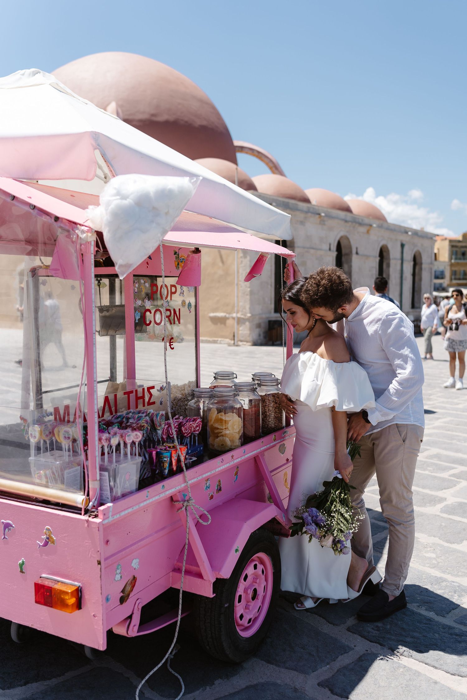 Wedding couple sharing a tender moment beside a charming pink popcorn cart in a Greek town square.