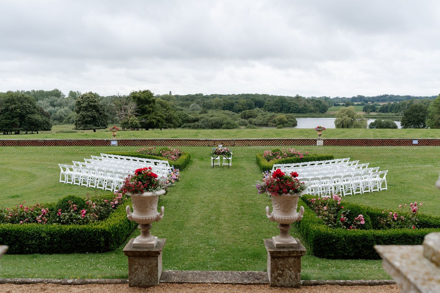 Outdoor wedding ceremony setup with white chairs on manicured lawn overlooking scenic lake and gardens.