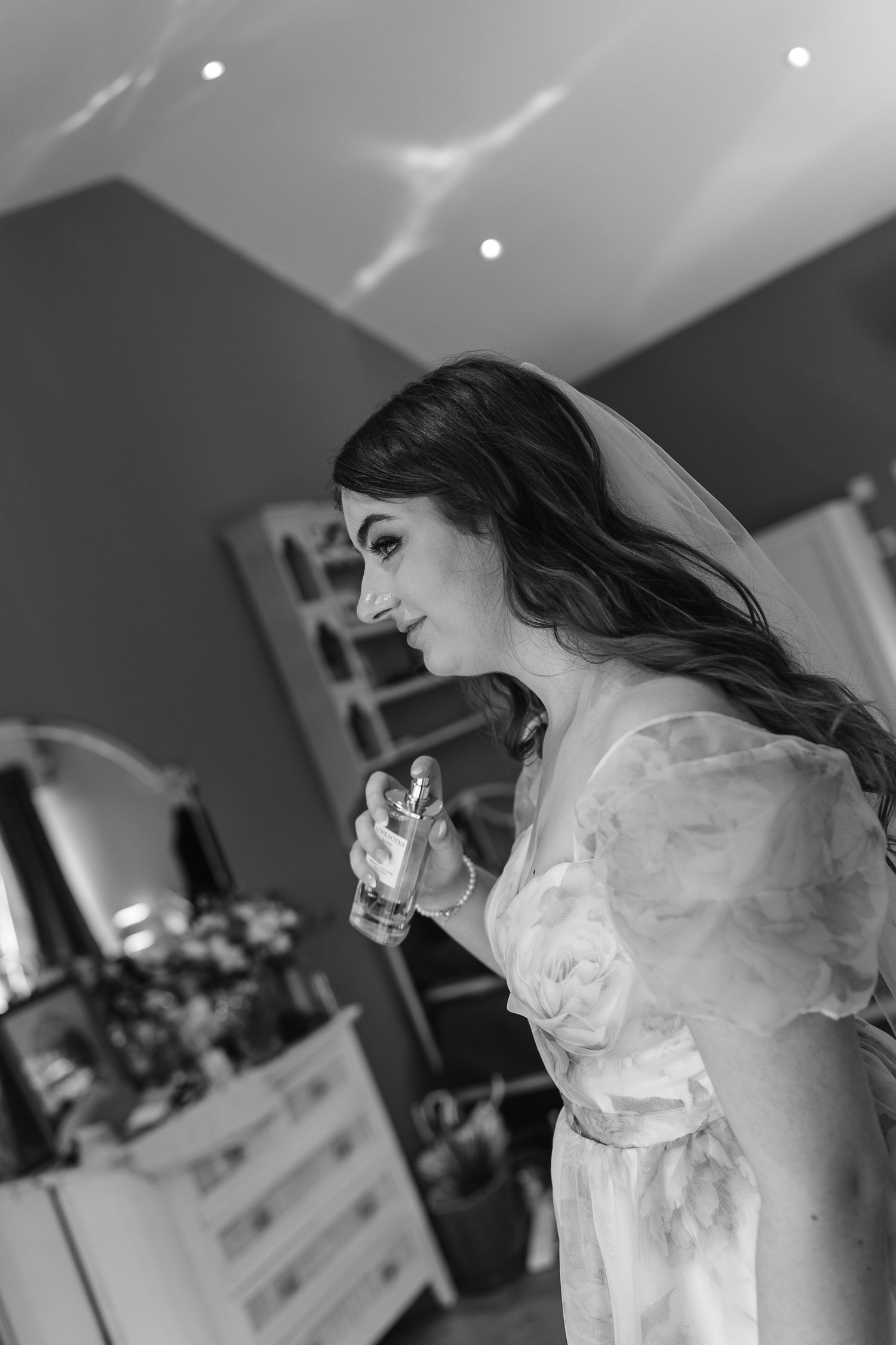 Black and white photo of a bride in a floral dress applying perfume while getting ready on her wedding day.