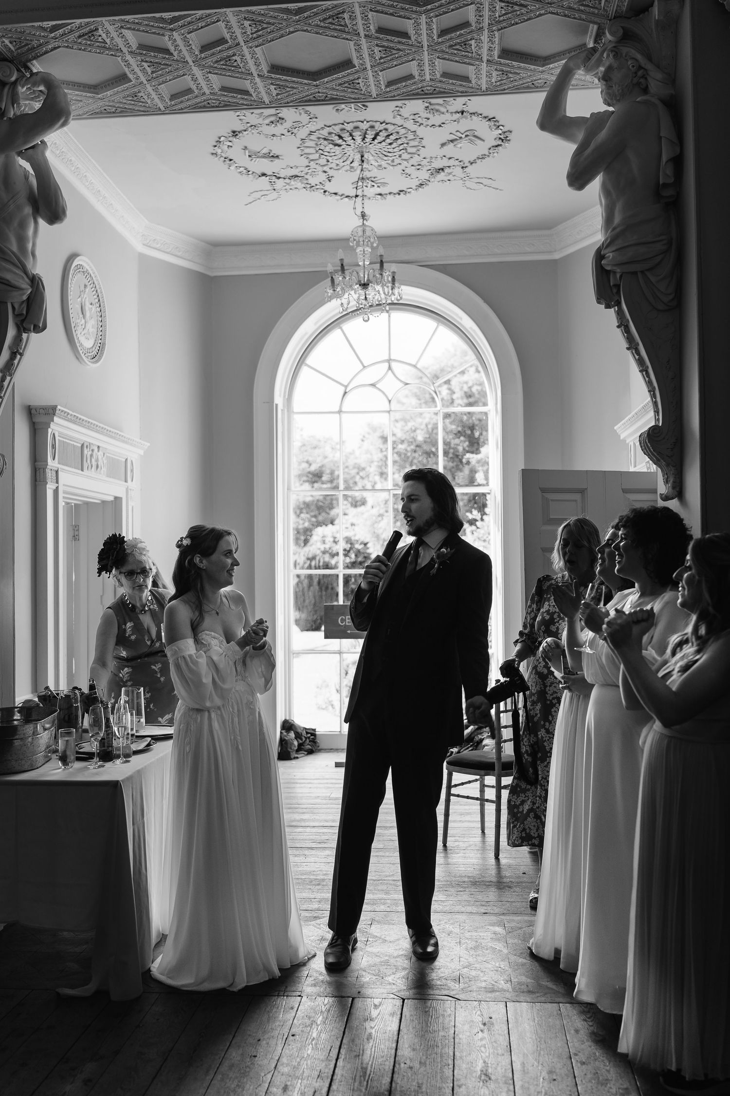 Groom gives speech in ornate room as bride and guests applaud in black and white photo.