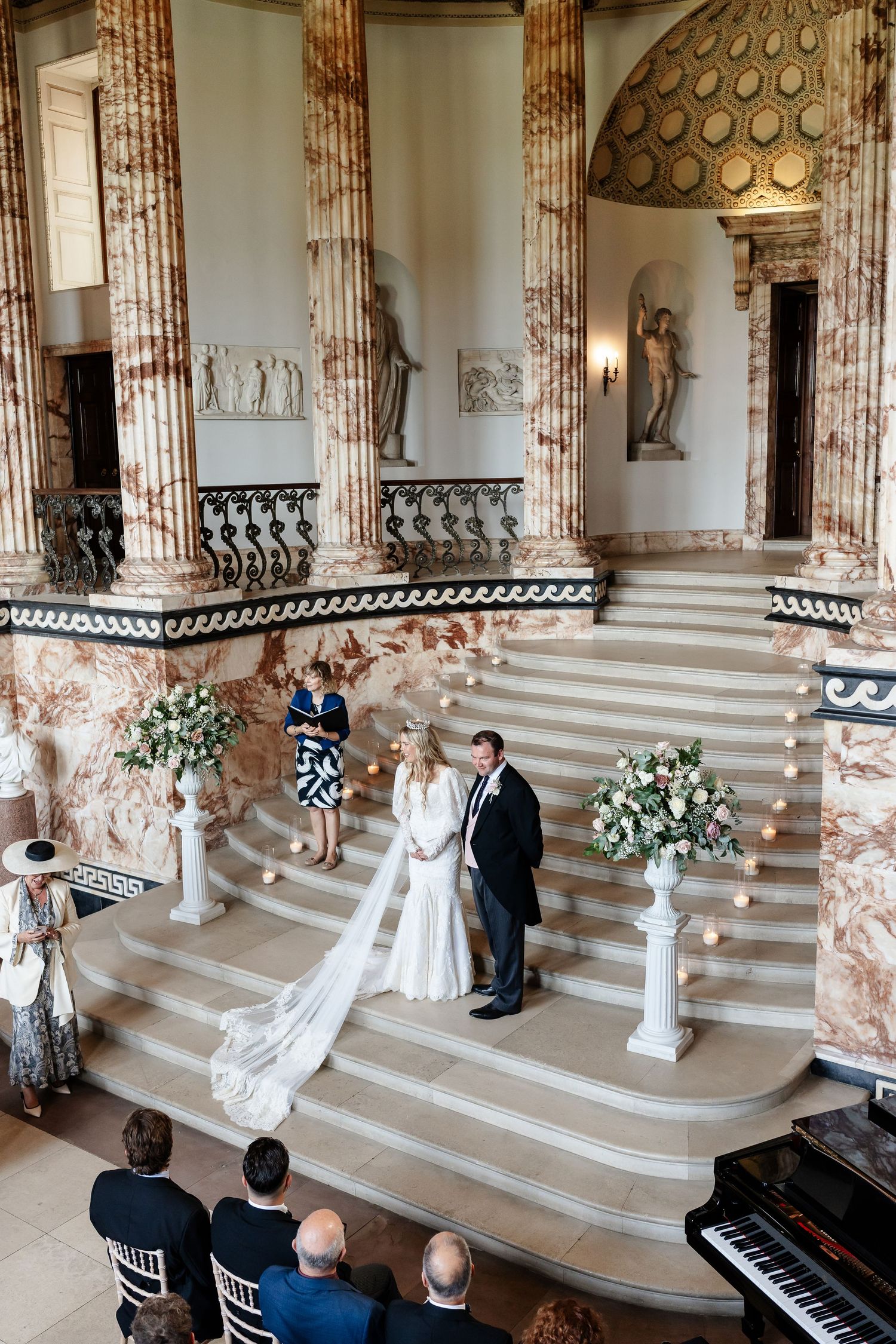 Bride and groom exchange vows on grand marble staircase in elegant neoclassical venue with floral arrangements.