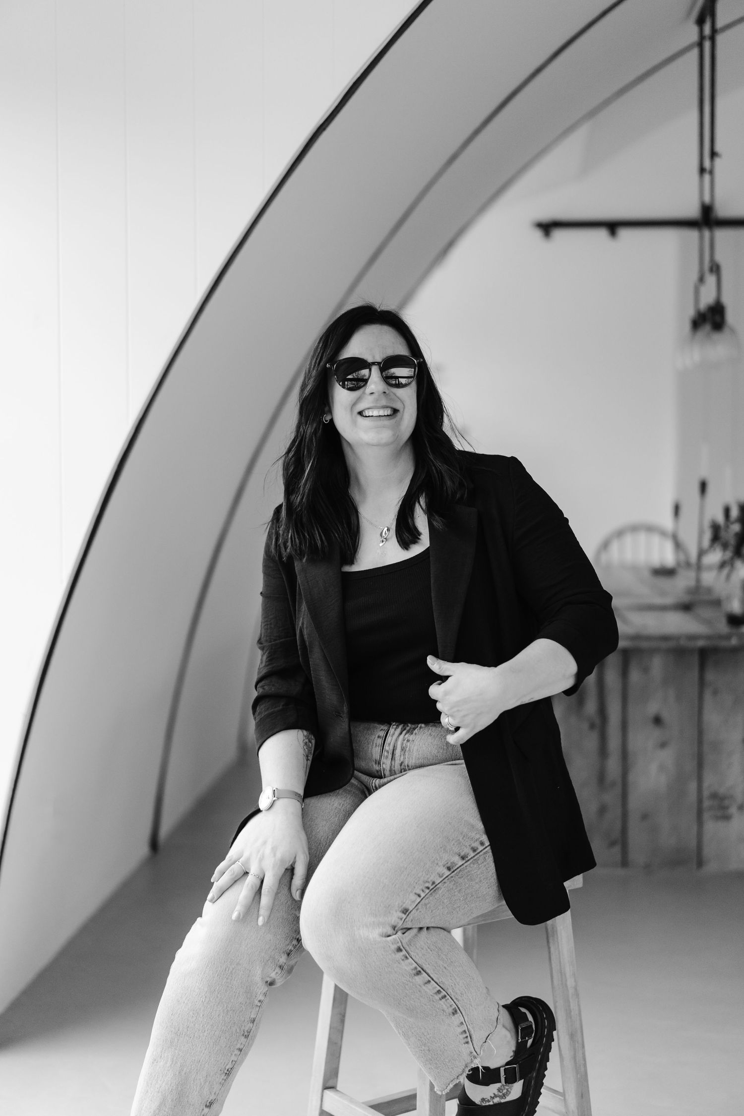 Norfolk Wedding Photograher Cherrelle laughing  in black blazer and sunglasses sitting on a stool and smiling in a modern branding studio