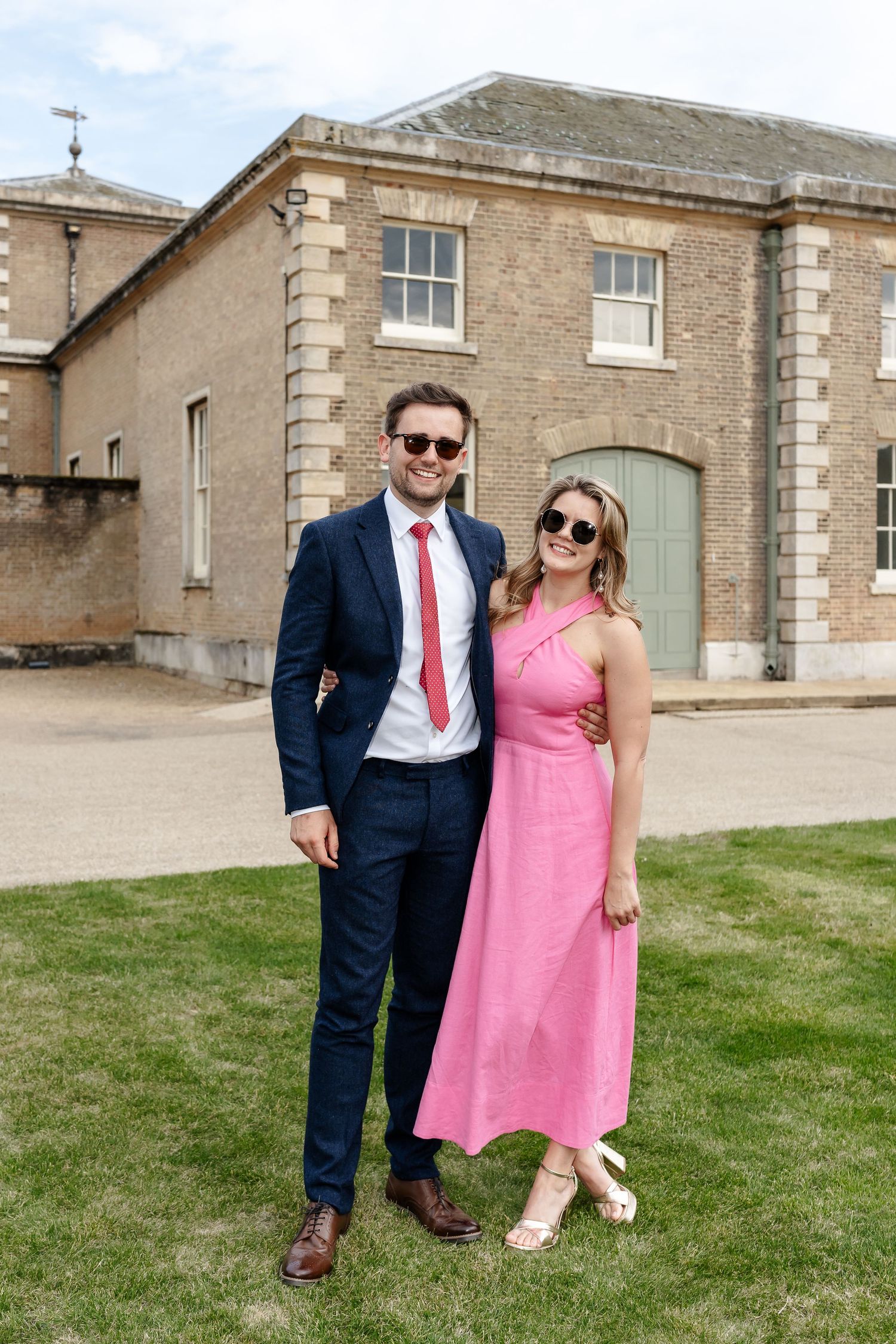 Couple poses outdoors at wedding venue, man in navy suit, woman in pink dress, both wearing sunglasses.
