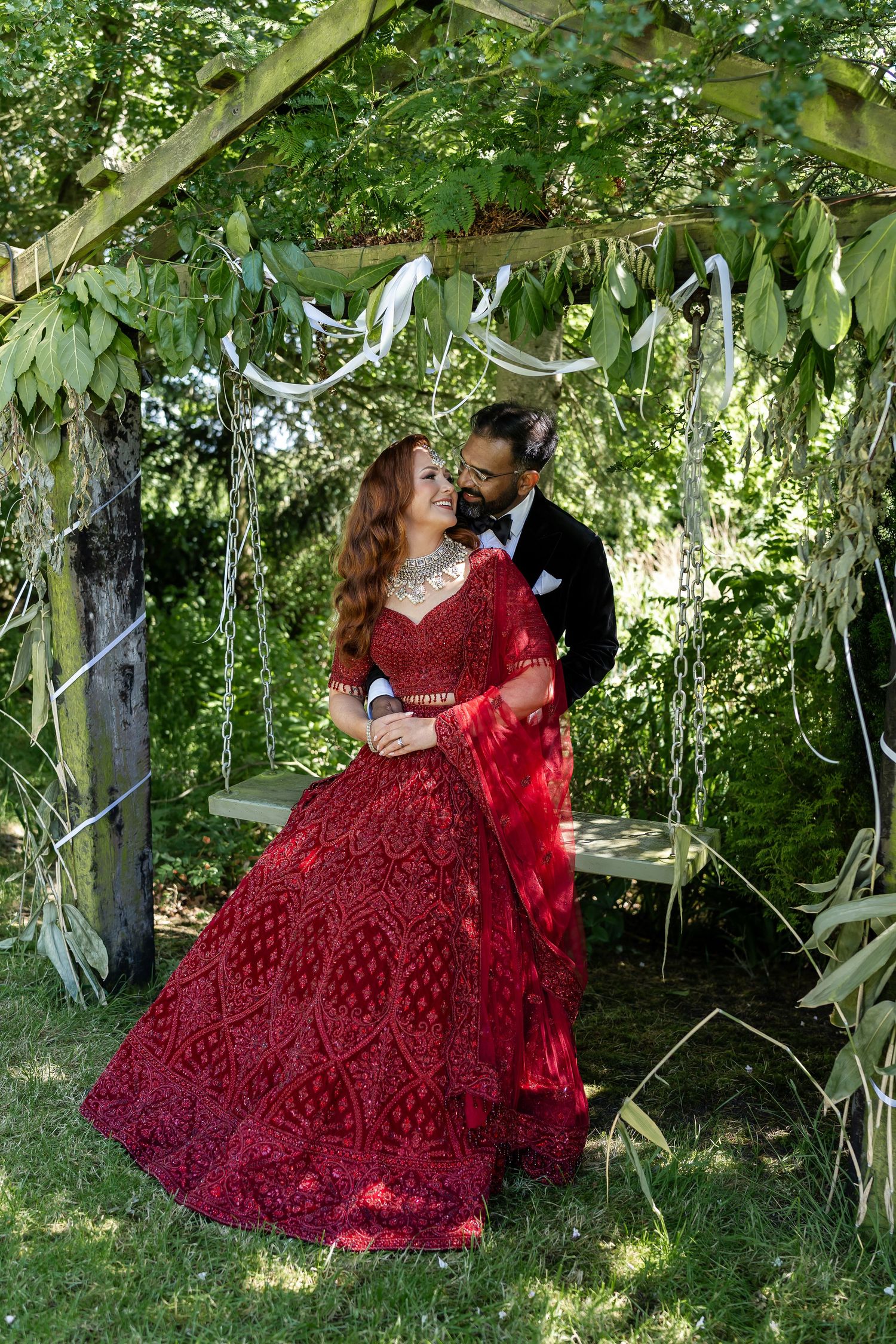 Couple in elegant red bridal lehenga and black suit embrace under leafy garden pergola.
