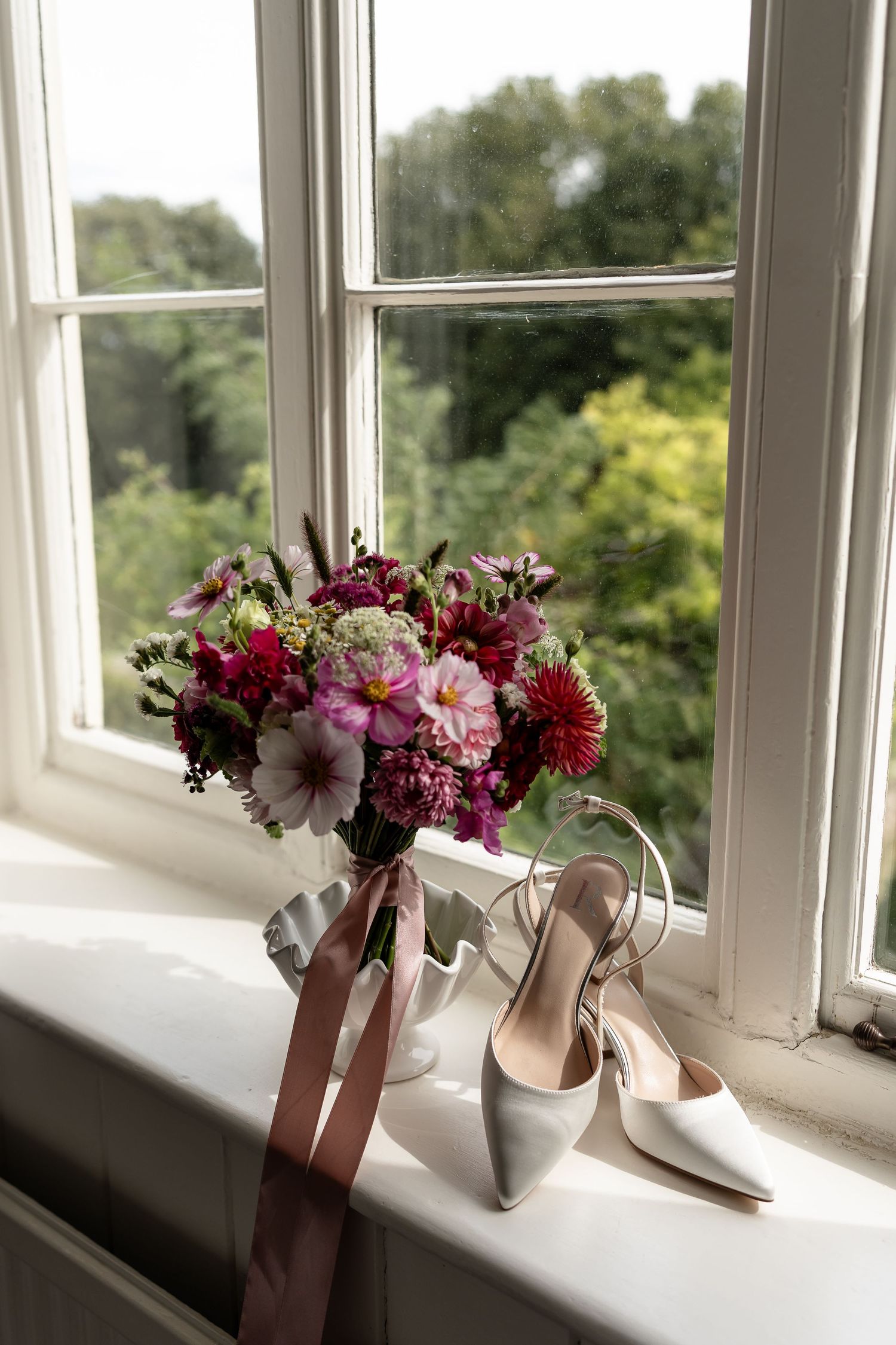 Bridal bouquet of pink and red flowers with satin ribbon beside white heels on a windowsill.