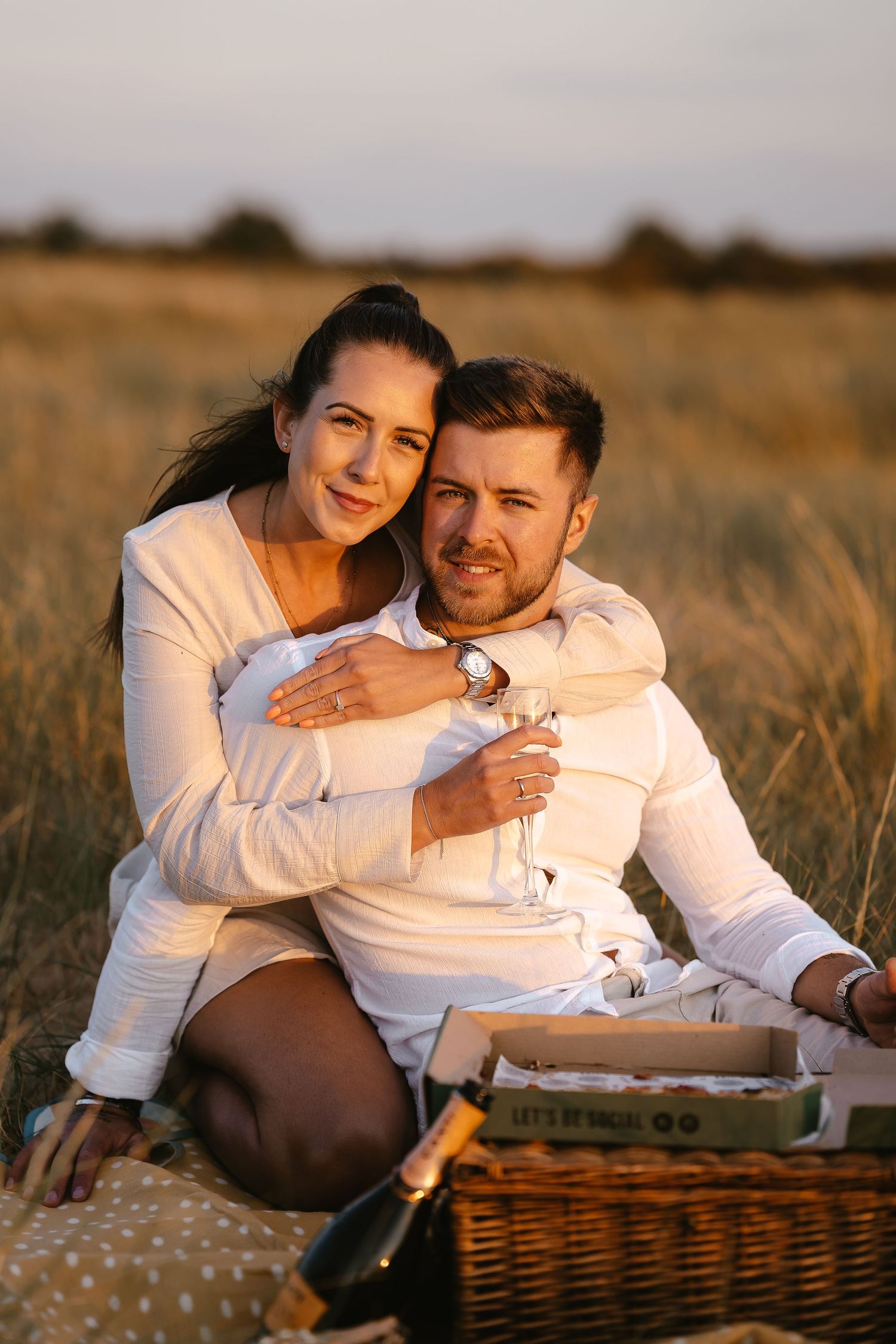 Smiling couple in matching white outfits sharing a romantic picnic with champagne in a sunlit field.