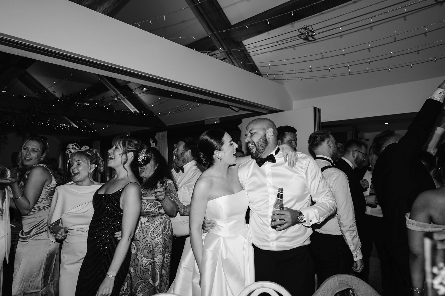Bride and groom share a kiss on a crowded wedding reception dance floor in black and white.