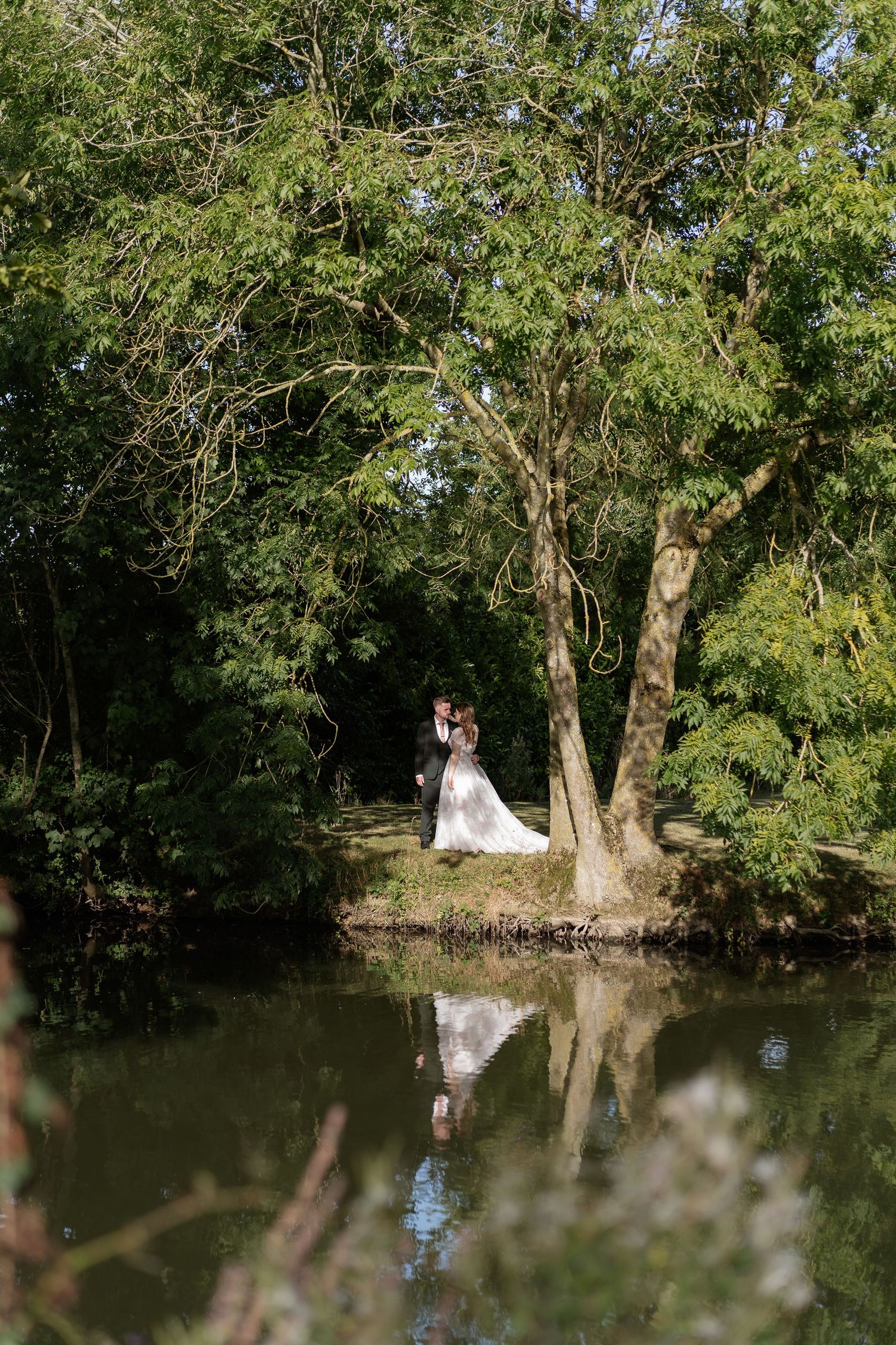 Bride and groom embrace by riverside tree with their reflection mirrored in calm water below.