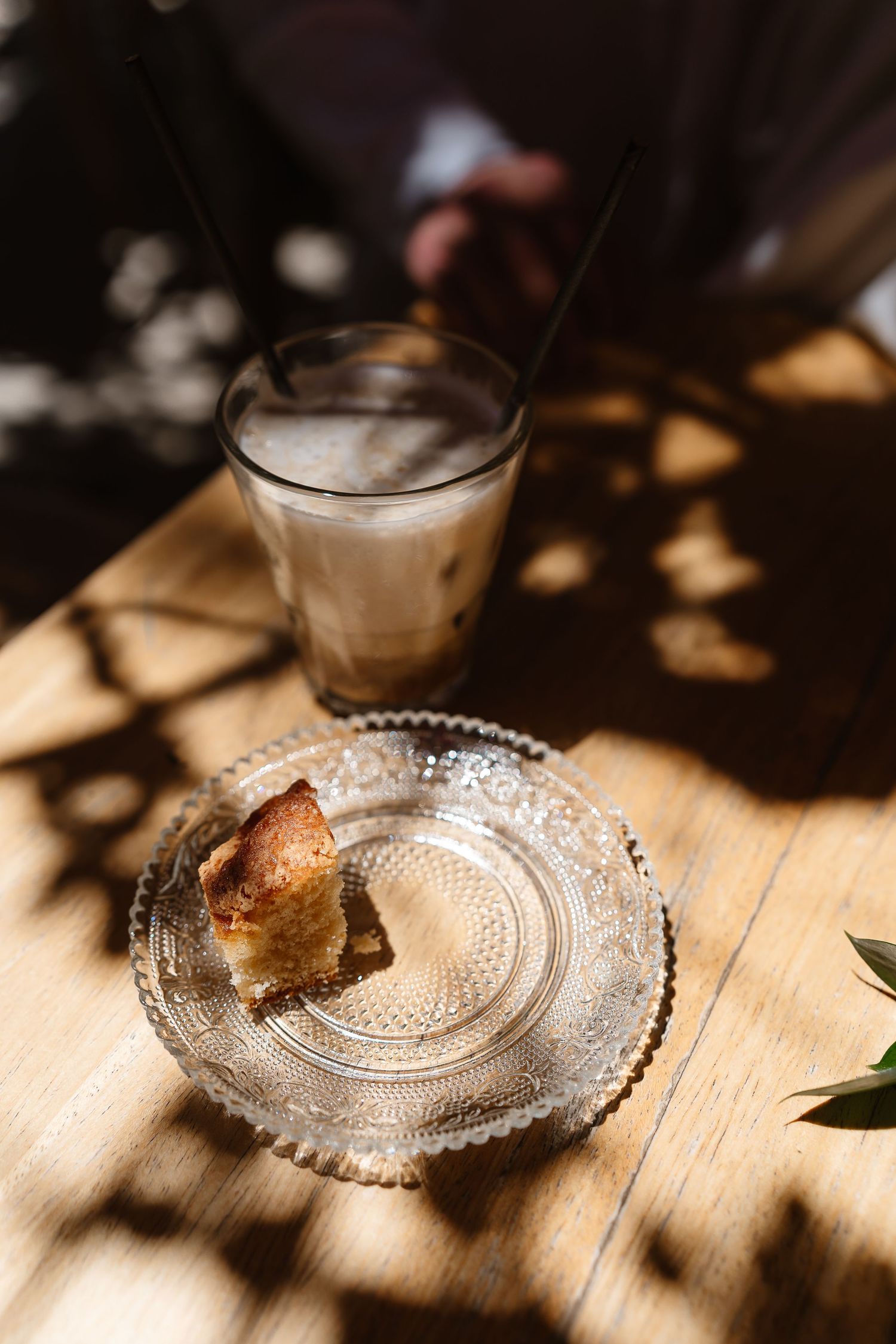 Iced latte and pastry on a glass plate on a sunlit wooden table with dappled shadows.