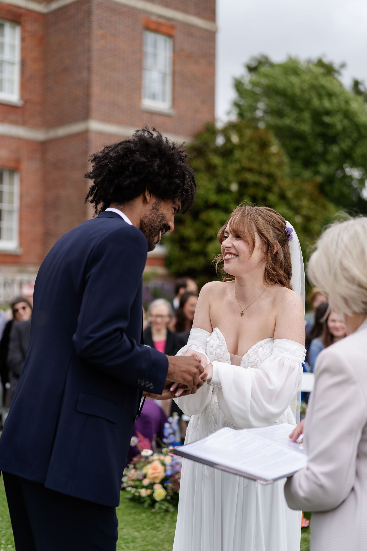 Bride in white gown smiles as groom holds her hands during heartfelt outdoor wedding ceremony vow exchange.