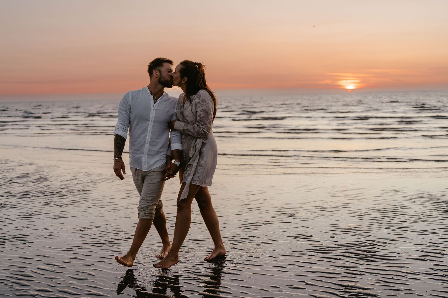 Couple sharing a kiss while walking barefoot on beach at vibrant pink and orange sunset.