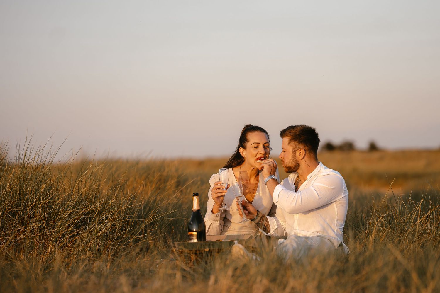Couple sharing food and champagne during a romantic picnic in a golden grassy field at dusk.