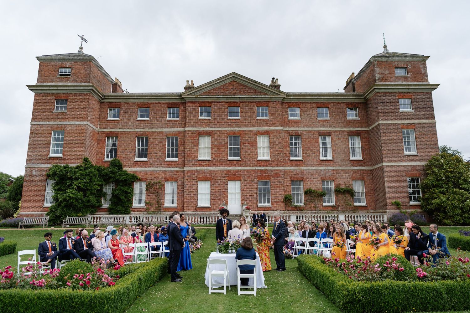 Wide shot of elegant outdoor wedding ceremony with guests seated on lawn of grand red brick manor.