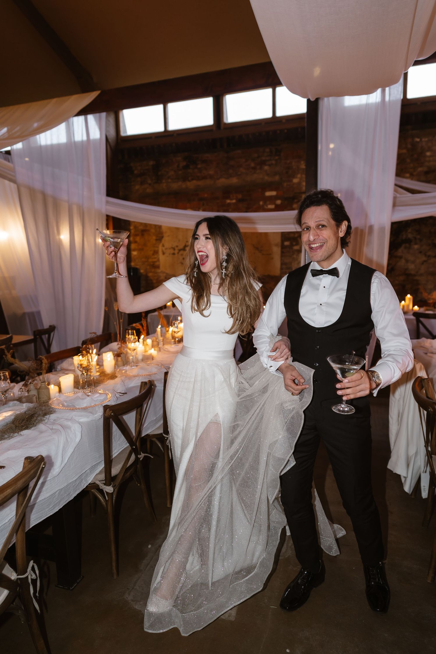Joyful bride and groom celebrating at candlelit rustic barn wedding reception with draped fabric ceiling.