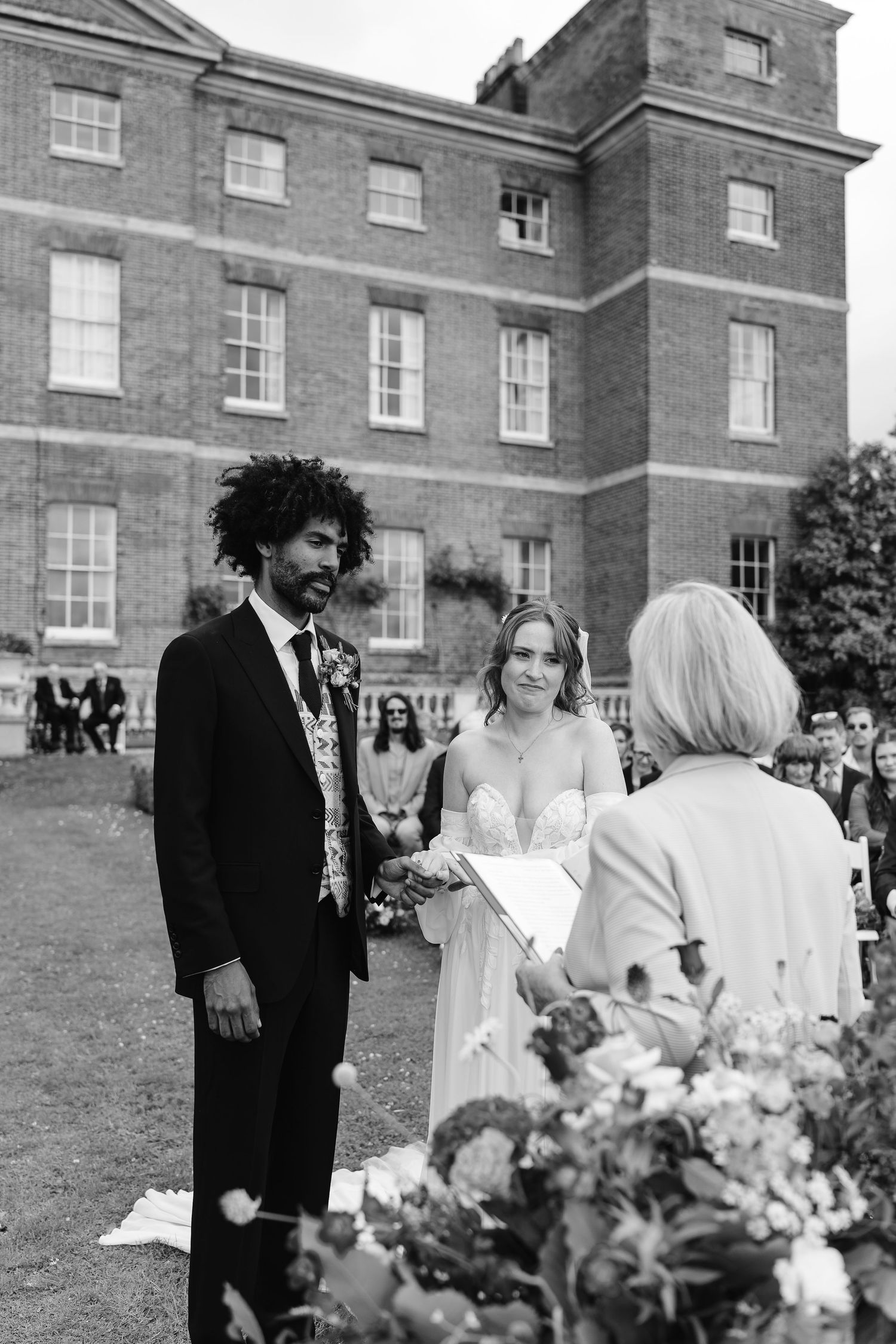 Black and white photo of bride and groom exchanging vows outdoors before a grand brick manor house.