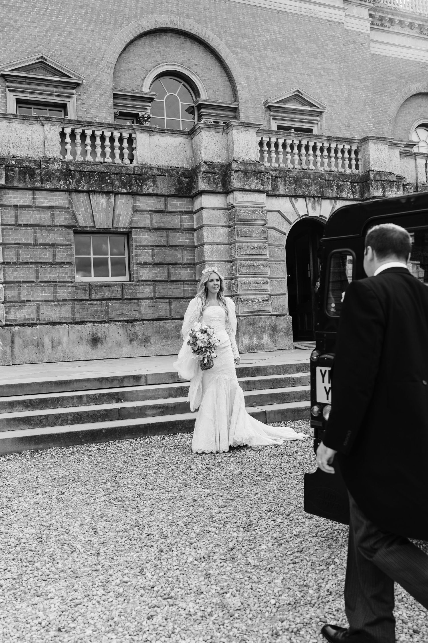 Black and white photo of bride in wedding gown holding bouquet outside grand historic estate building.